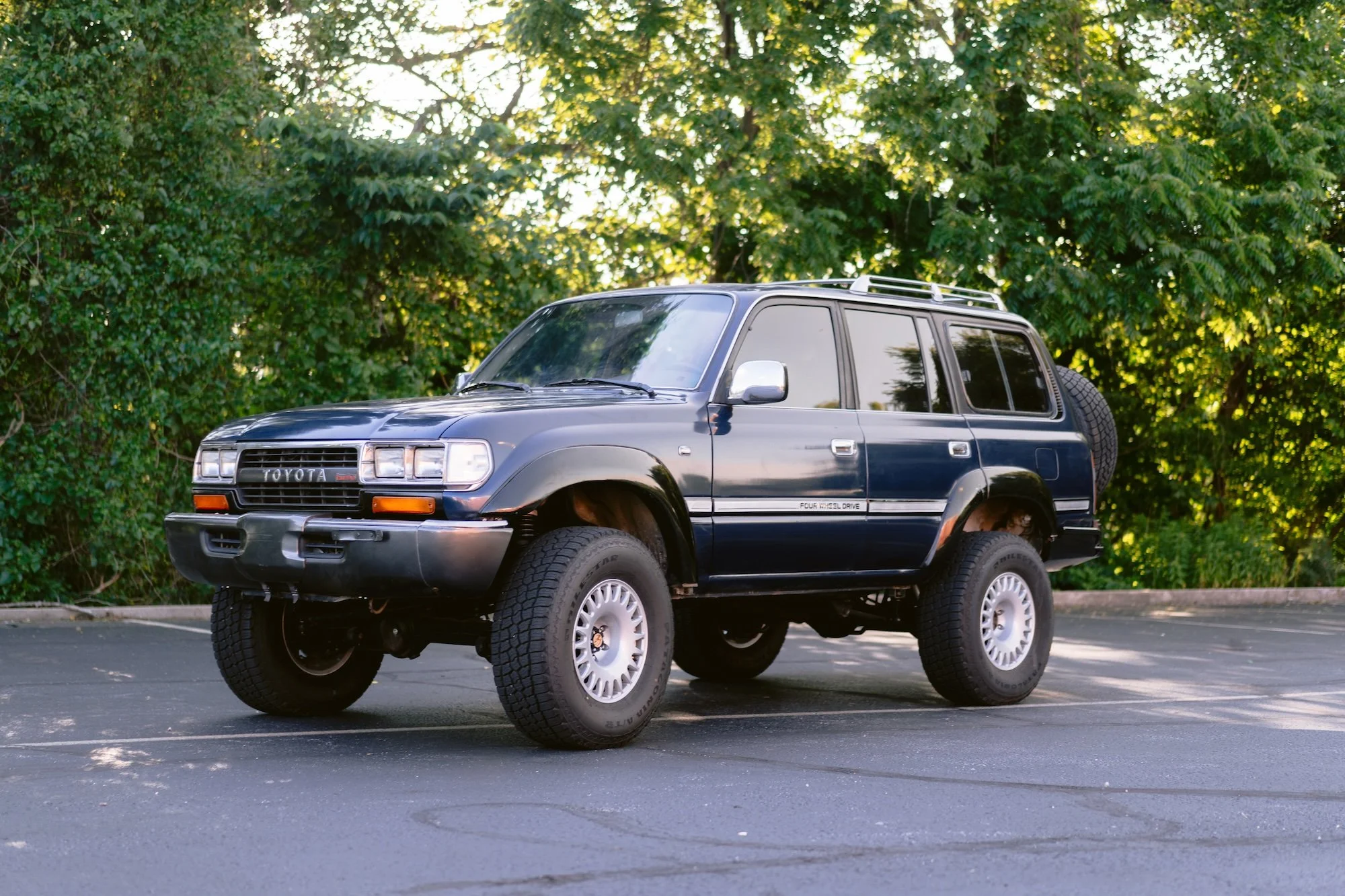 A black vintage Toyota 4Runner SUV with large tires and a spare tire mounted on the back, parked in an empty parking lot with green trees in the background.
