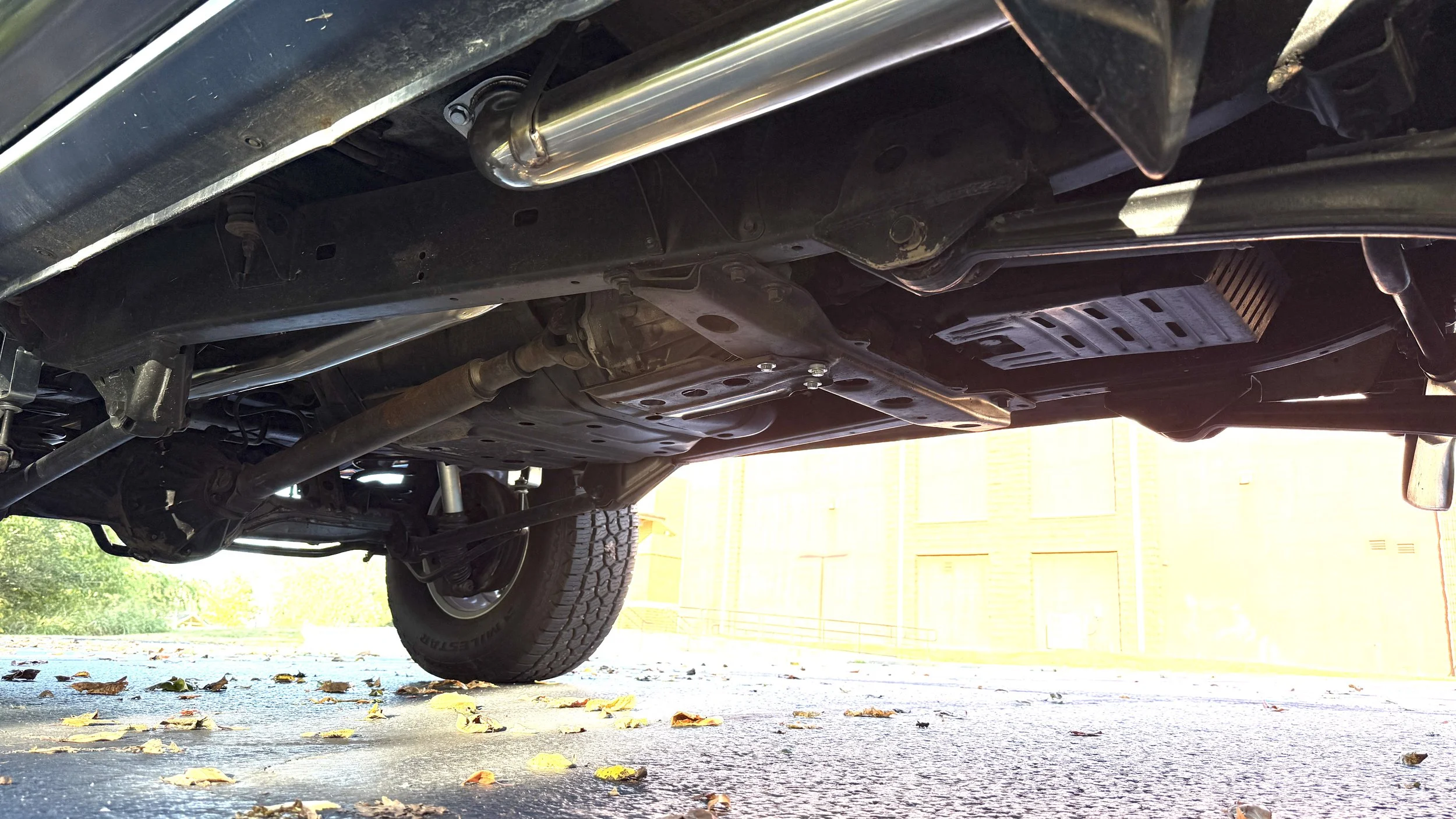 Underside of a vehicle showing exhaust pipe, suspension, and tire on a paved surface with fallen leaves and a yellow building in the background.
