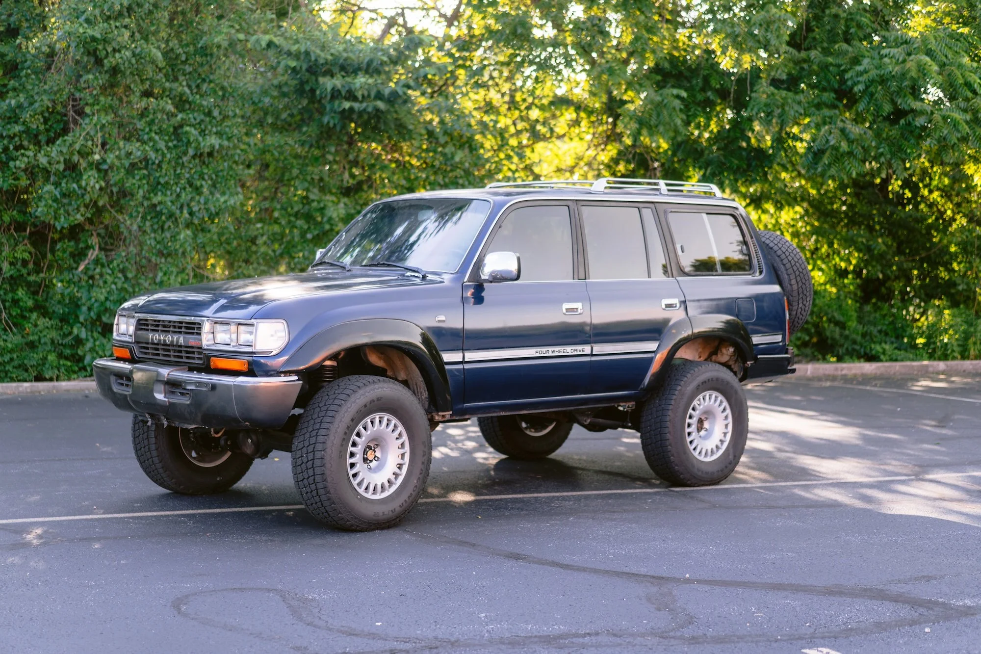 A black Toyota SUV with a missing front wheel parked in an outdoor lot surrounded by green trees.