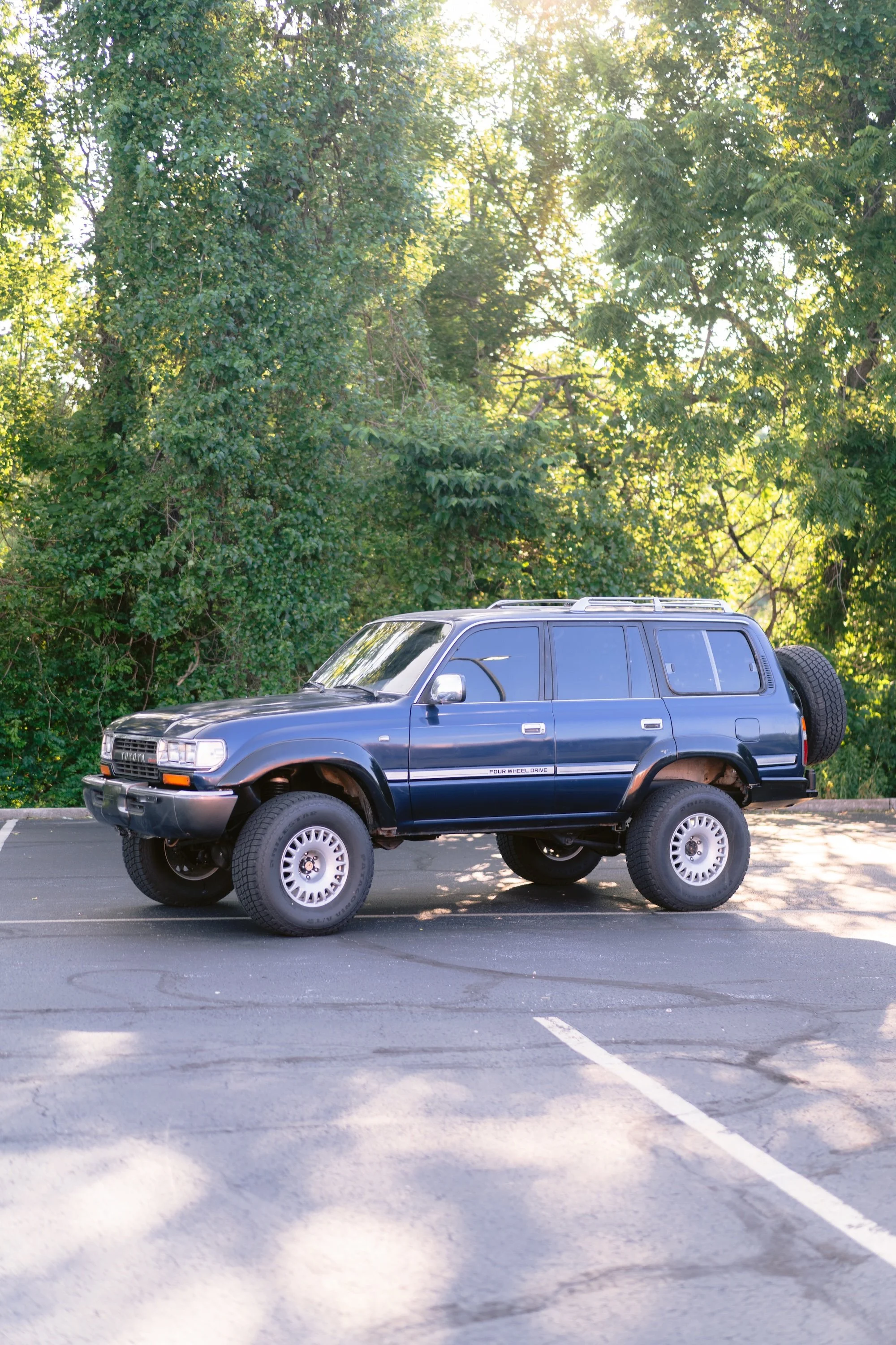 A blue SUV vehicle parked in an empty parking lot with a backdrop of green trees and sunlight filtering through the leaves.