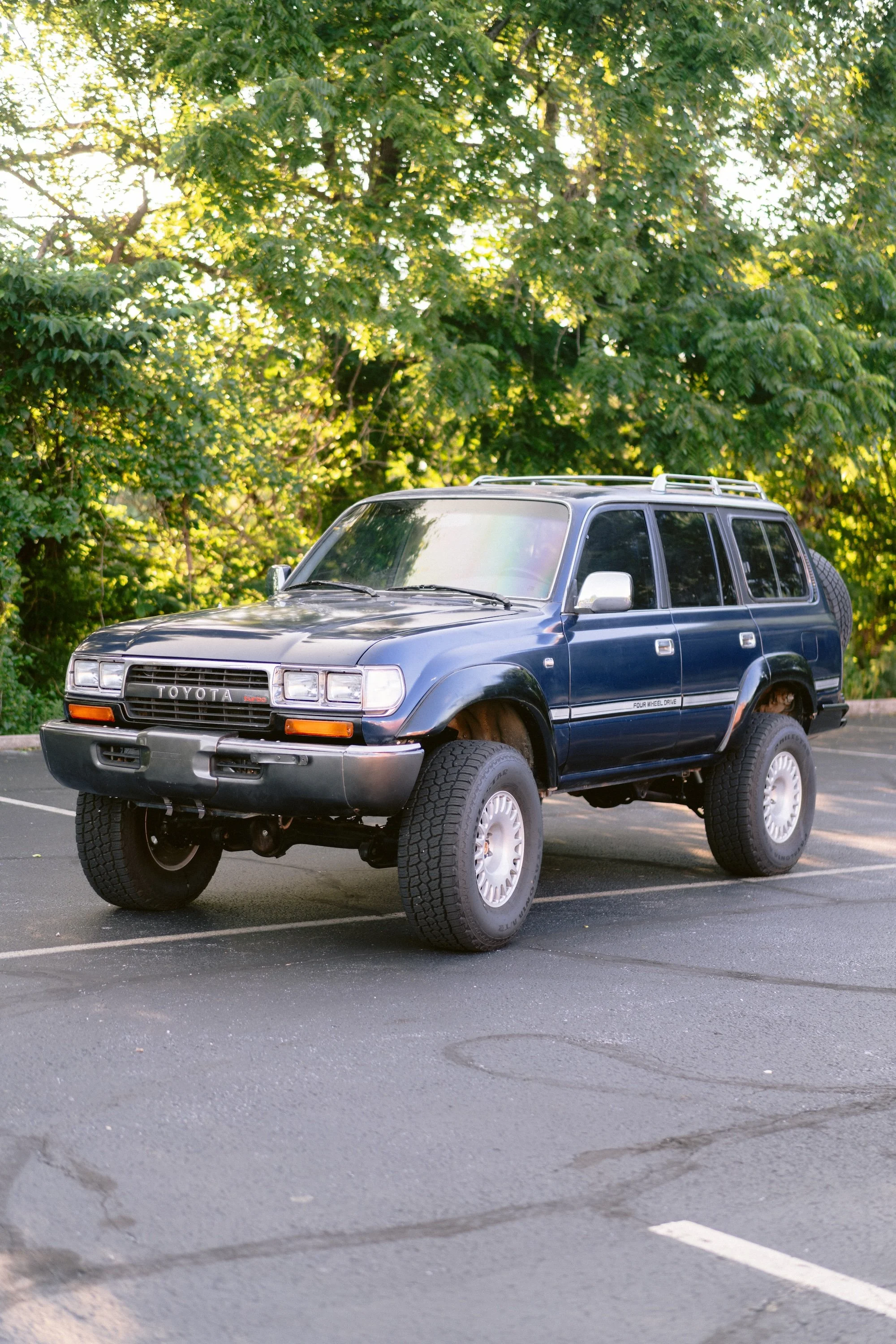 A blue Toyota 4Runner SUV parked in a parking lot with trees and green foliage in the background.