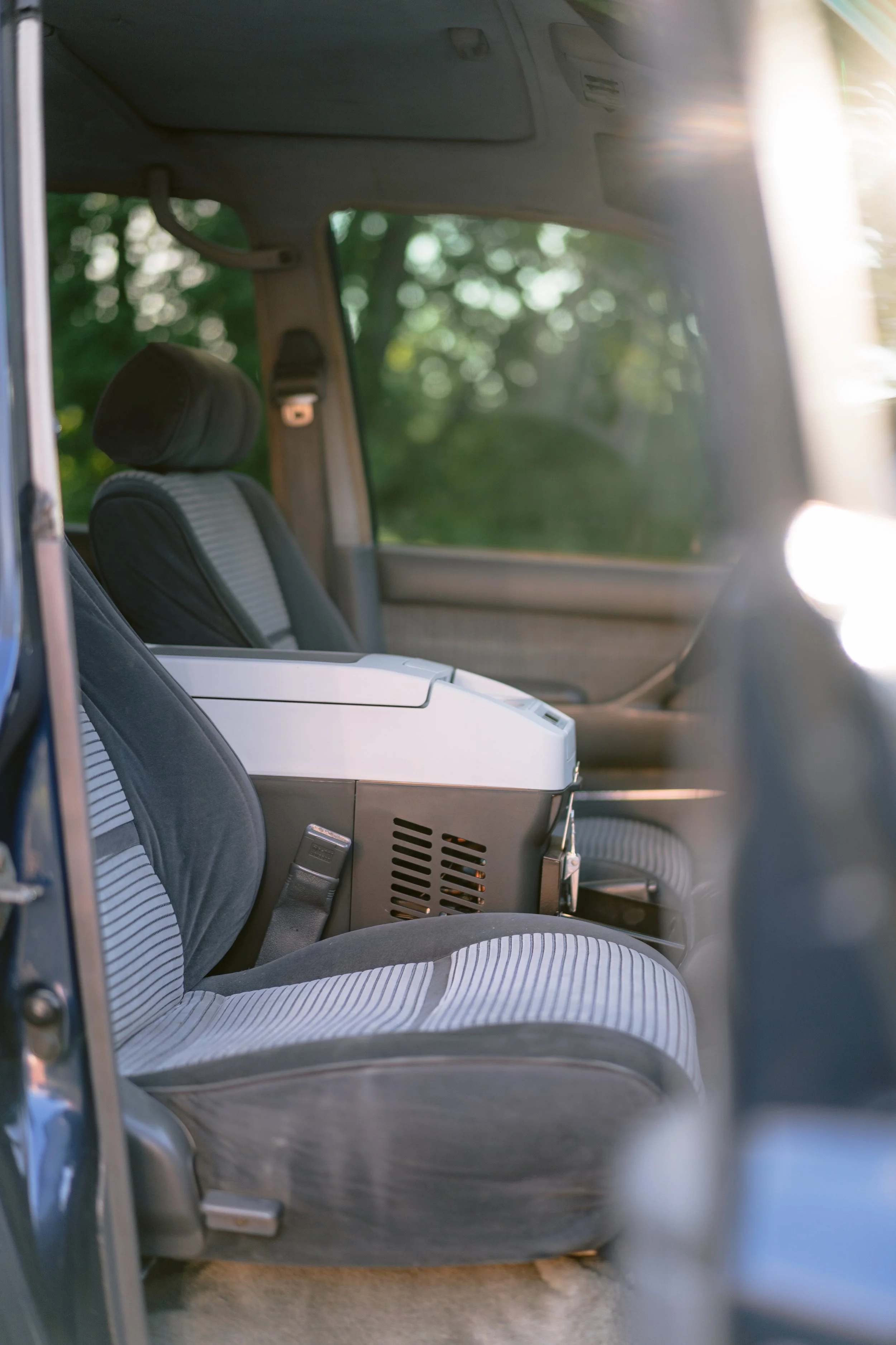 The interior of a vehicle showing the passenger seat, a white bag or small appliance on the seat, and a window with greenery outside, illuminated by sunlight.