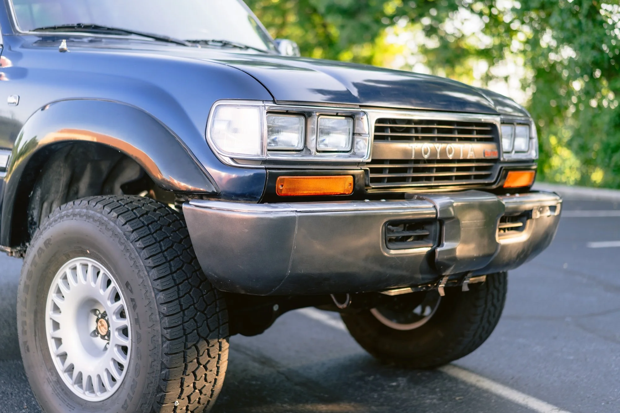 Close-up of a vintage black Toyota SUV's front end with headlights, grille, and tire visible, parked outdoors with a blurred green background.
