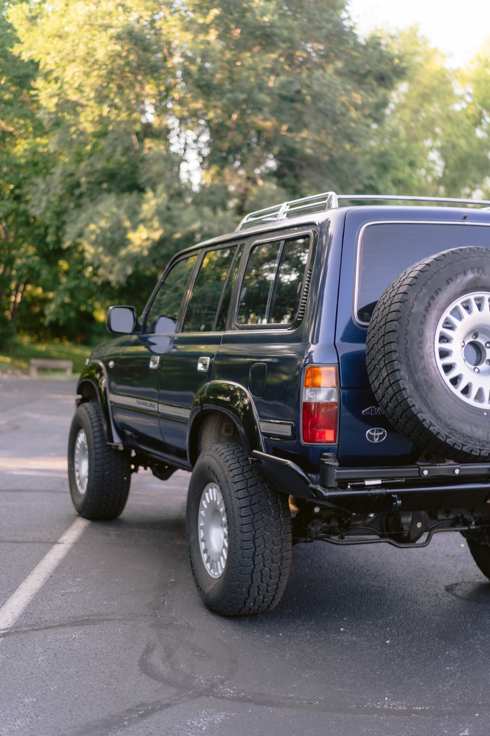 A blue Toyota SUV parked in a parking lot with trees in the background, a spare tire mounted on the rear, and sunlight filtering through the trees.