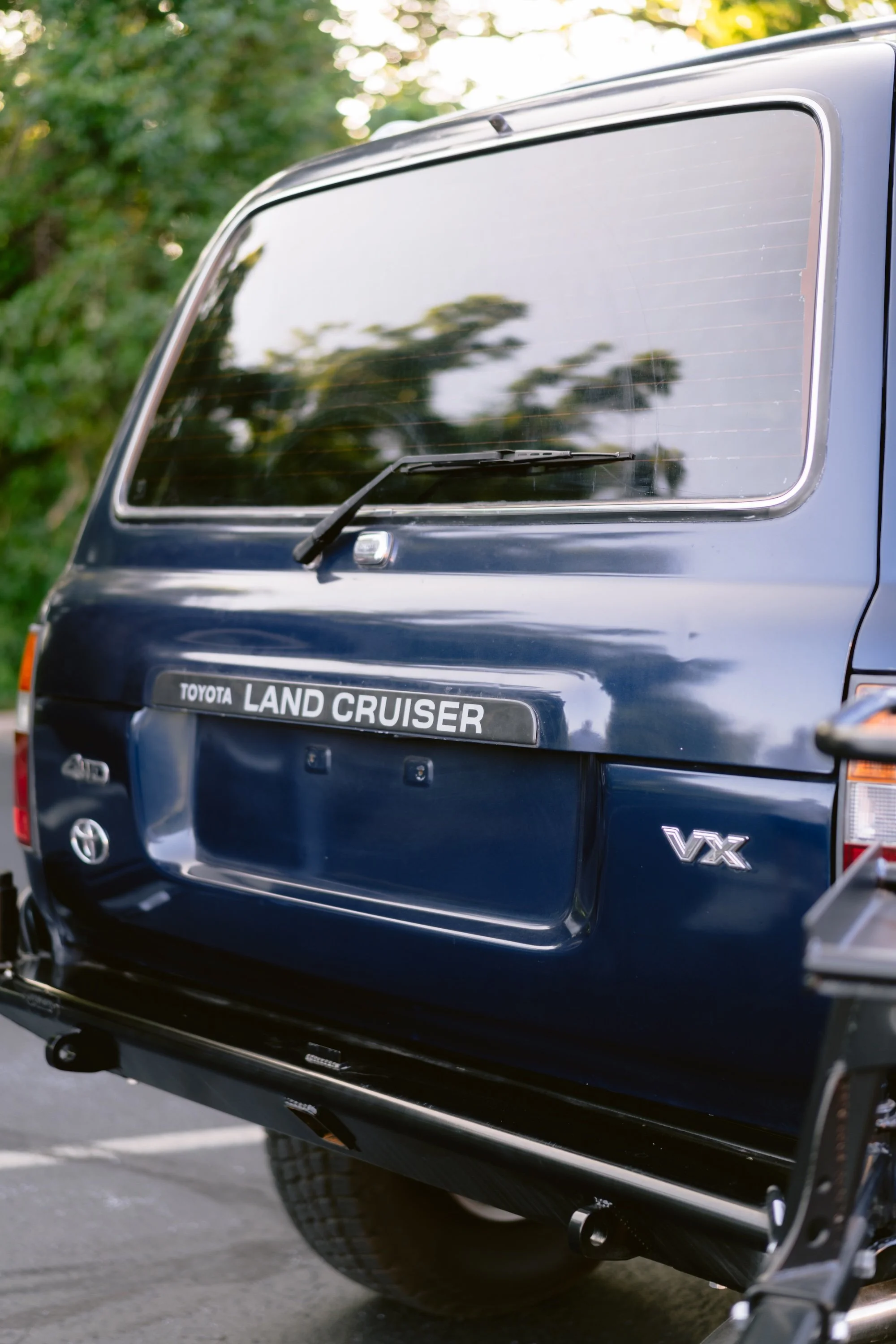 Close-up of a navy blue Toyota Land Cruiser V8 vehicle, showing the rear section with a tinted window, rear wiper, and the Land Cruiser badge, parked outdoors with green trees in the background.