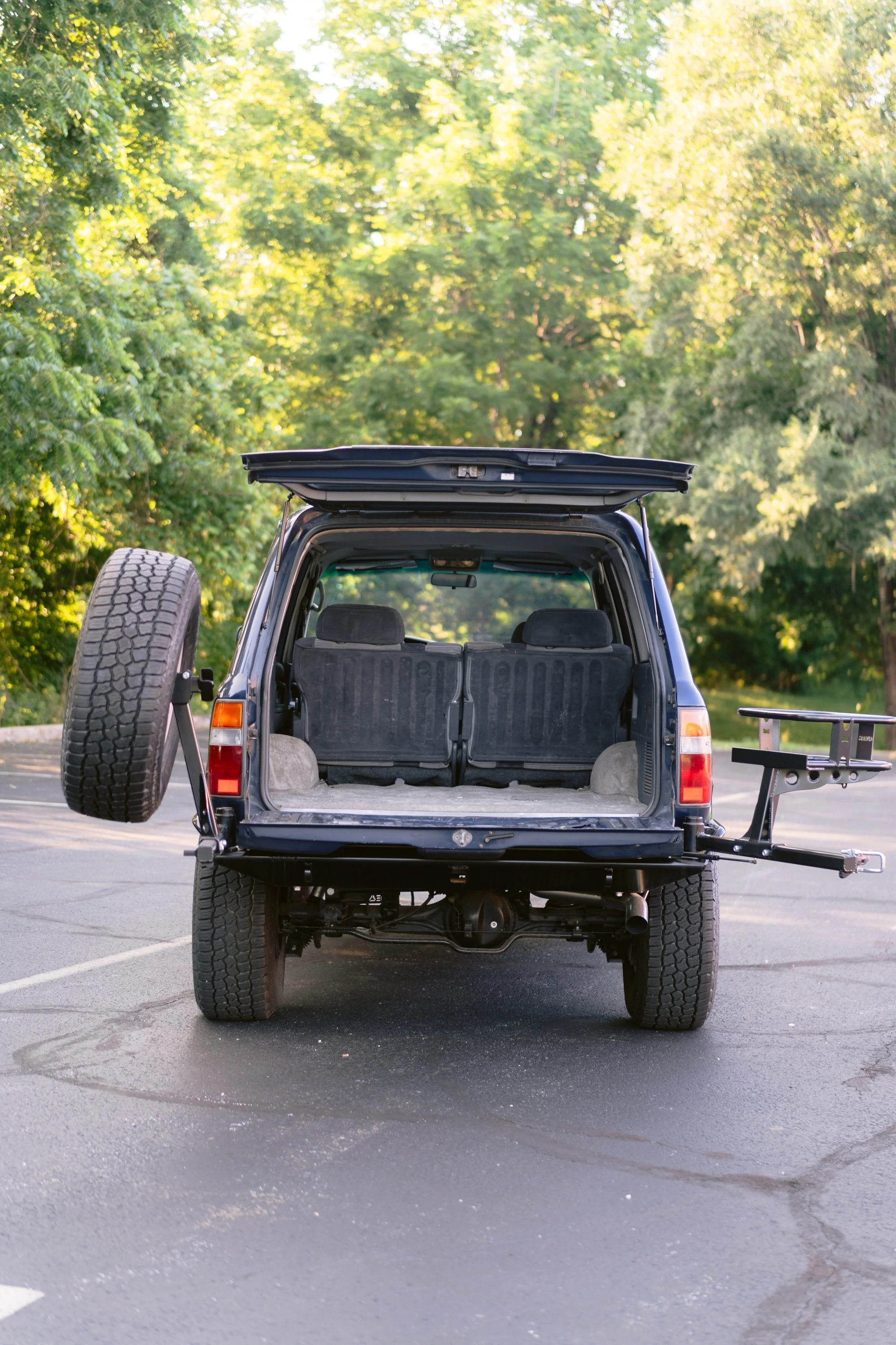 Rear view of a black SUV with an open trunk, a spare tire mounted on the left side, and green trees in the background.