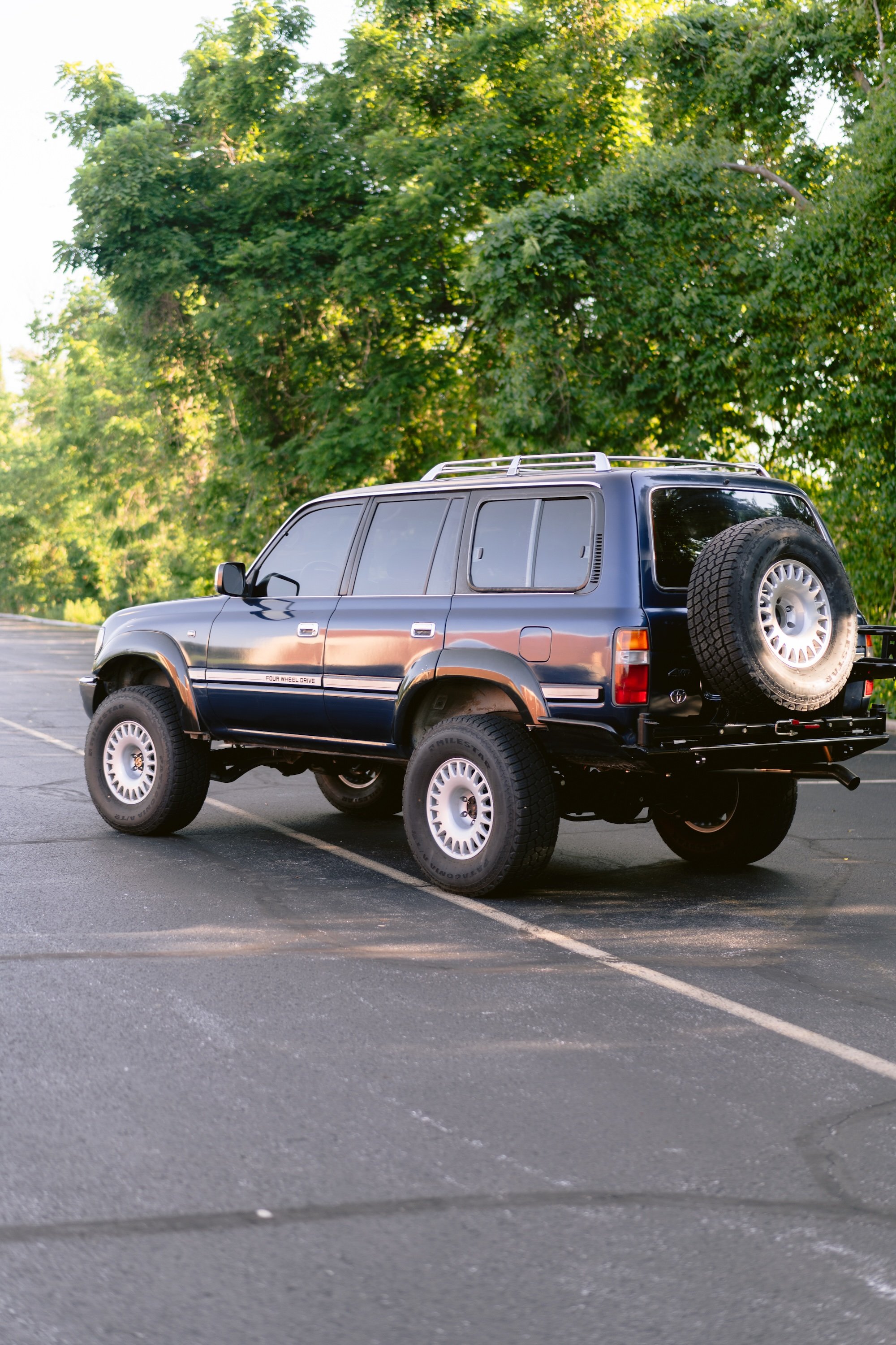 A dark blue SUV with a spare tire mounted on the back, parked in an empty parking lot with green trees in the background.