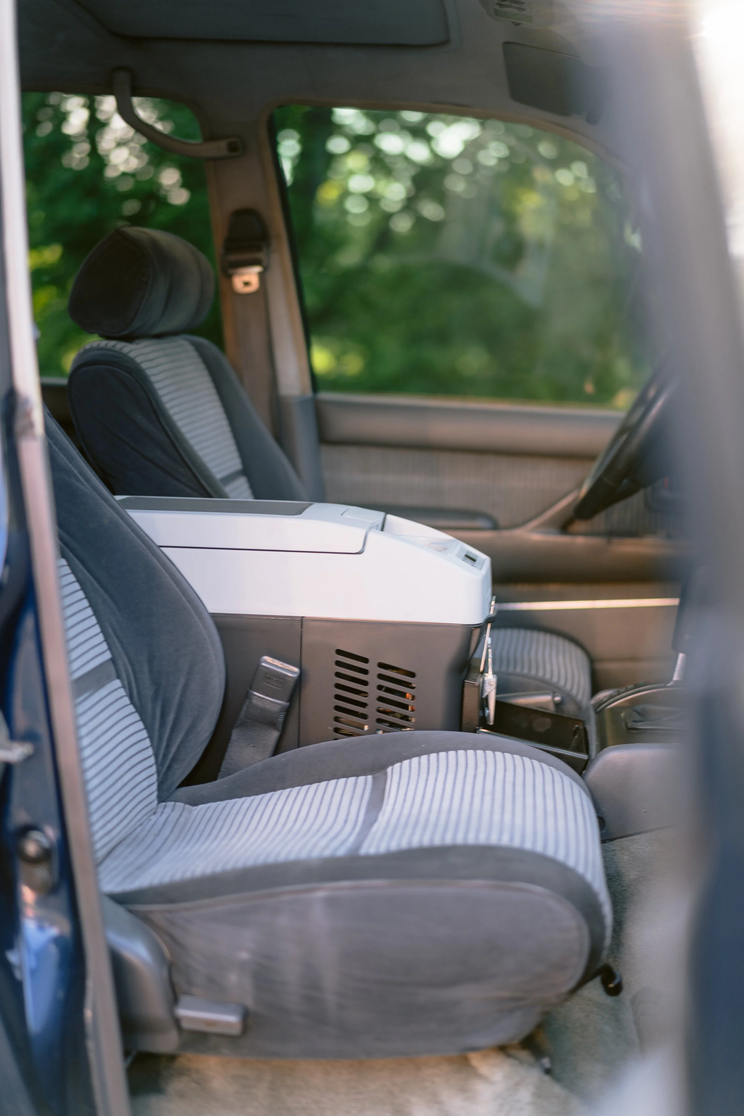 Interior of a vintage car with front seats having striped upholstery, and a white rectangular appliance or box placed near the passenger seat. The background shows blurred greenery outside the window.