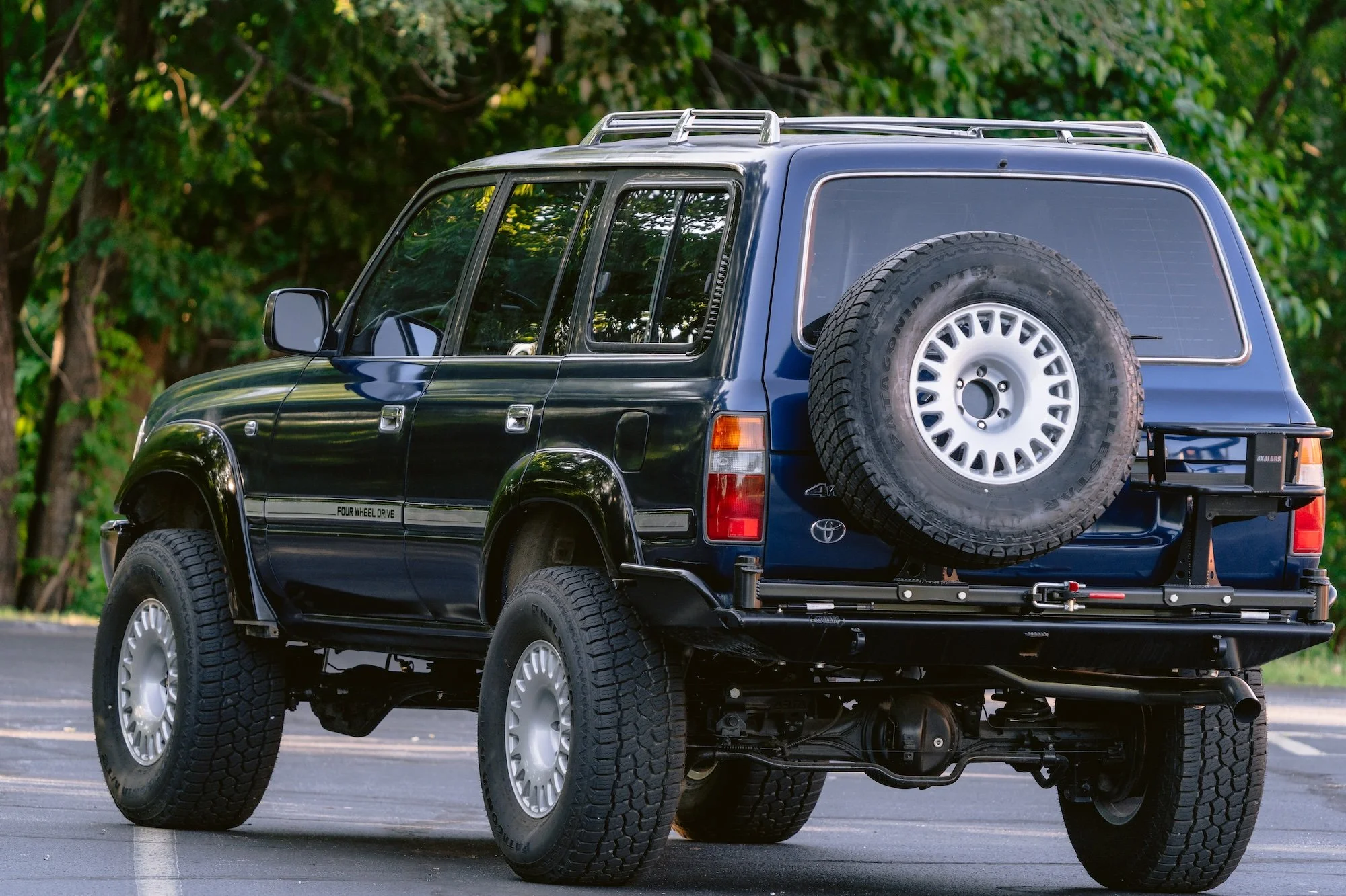 Back view of a black Toyota SUV with a full-sized spare tire mounted on the rear, parked in an outdoor lot with trees in the background.