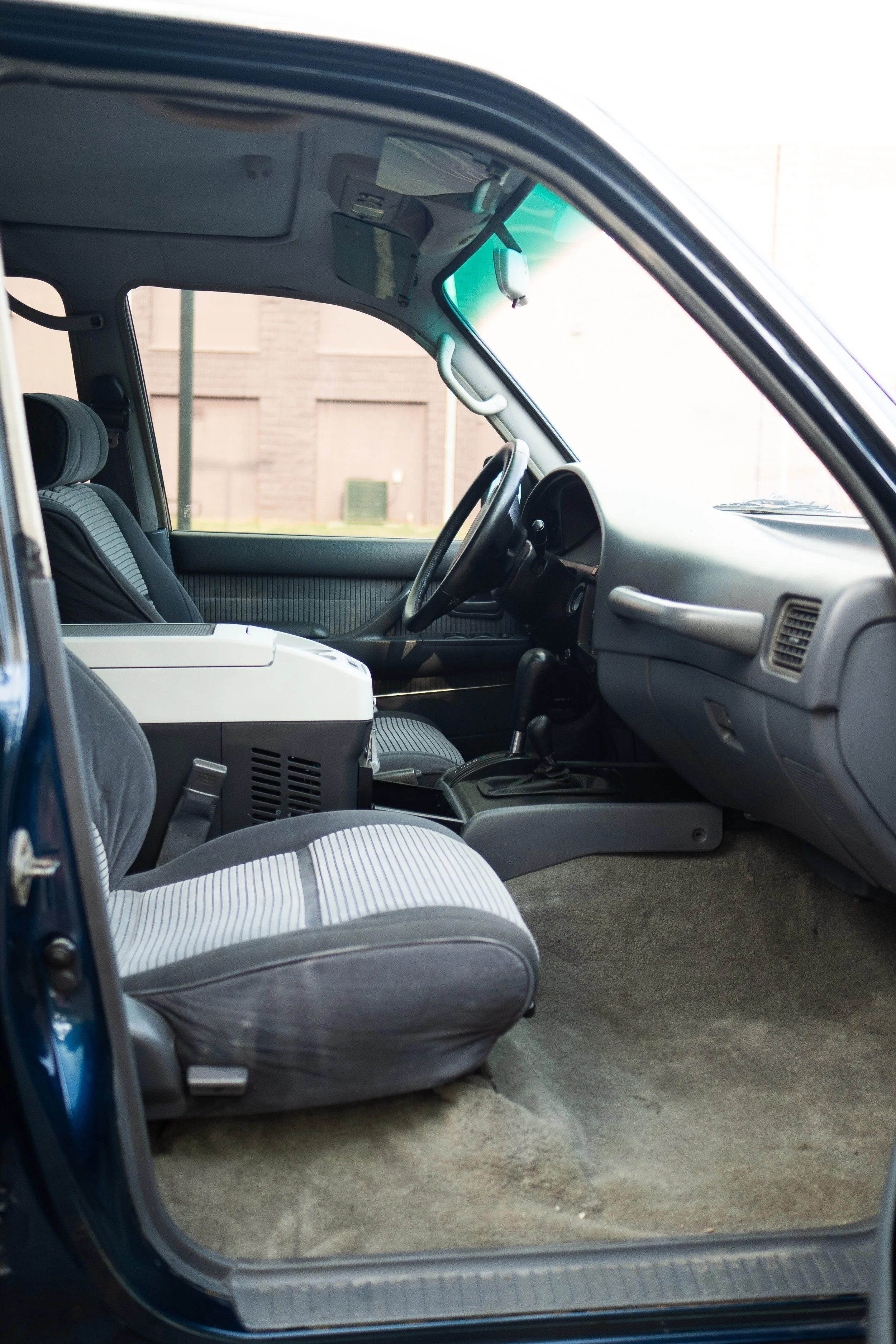 Interior view of an older model vehicle with striped fabric seats, a gray dashboard, and a steering wheel. An electronic device is visible on the passenger seat, and sunlight is illuminating the scene through the window.