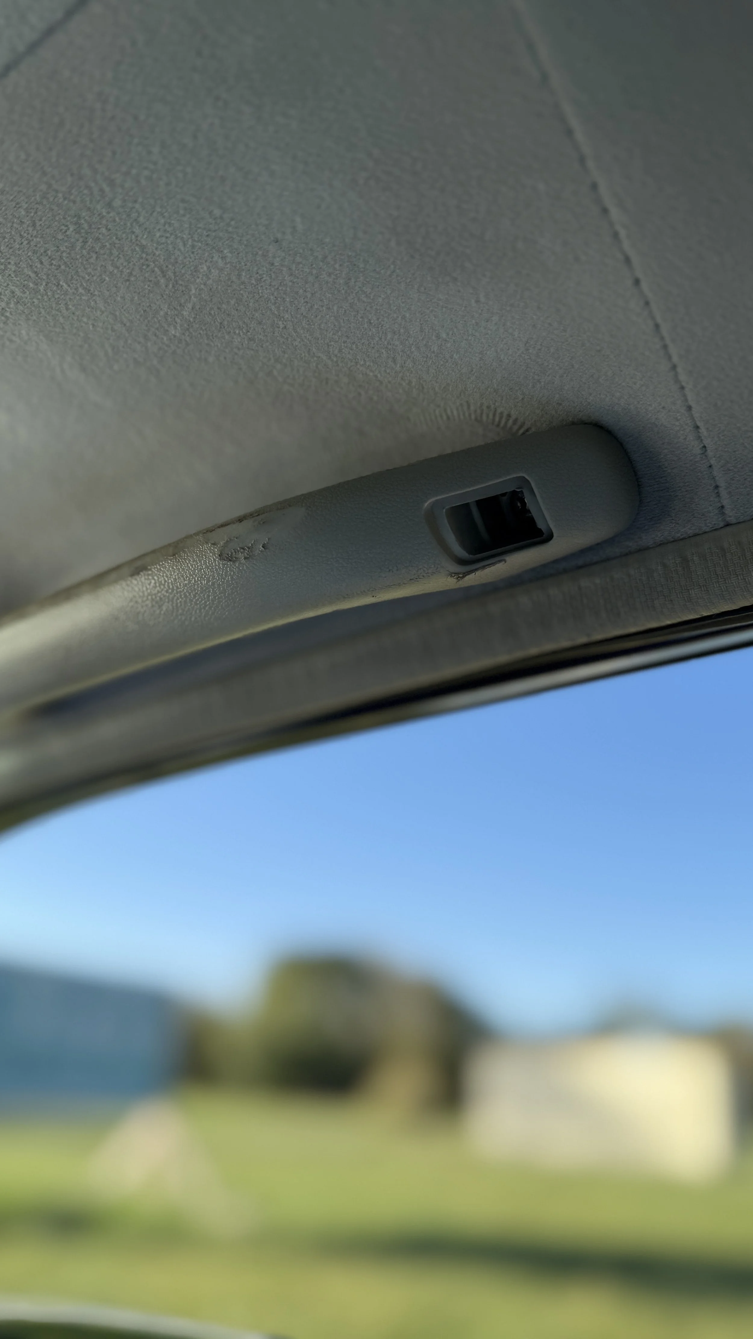 Close-up of the interior roof of a vehicle with a sunroof or moonroof, showing the control switch and fabric lining. The sky is visible through the open sunroof.