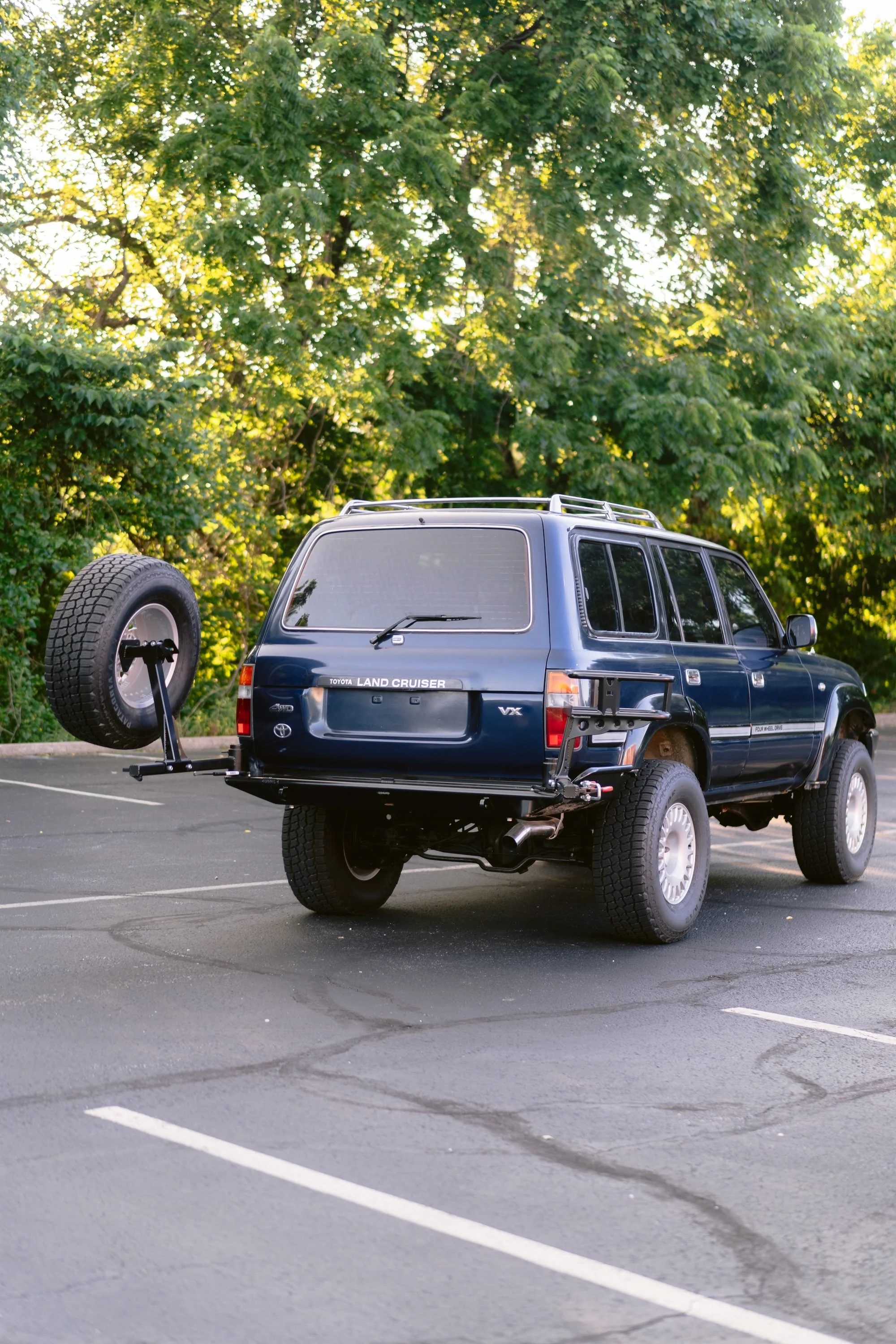 Blue Toyota Land Cruiser parked in an empty parking lot with a spare tire mounted on the back, trees in the background.