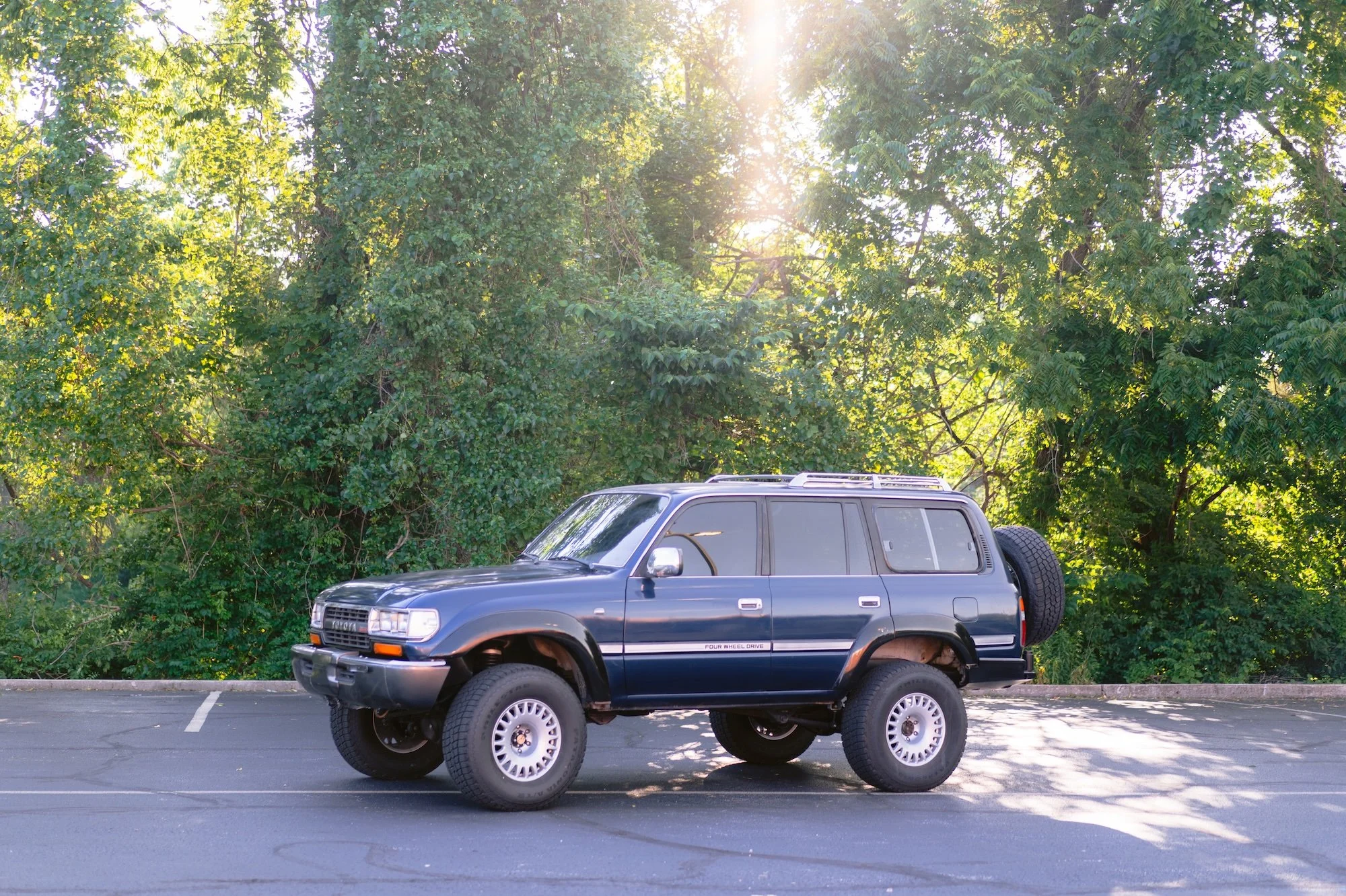 A black SUV parked in an empty parking lot with green trees and sunlight in the background.