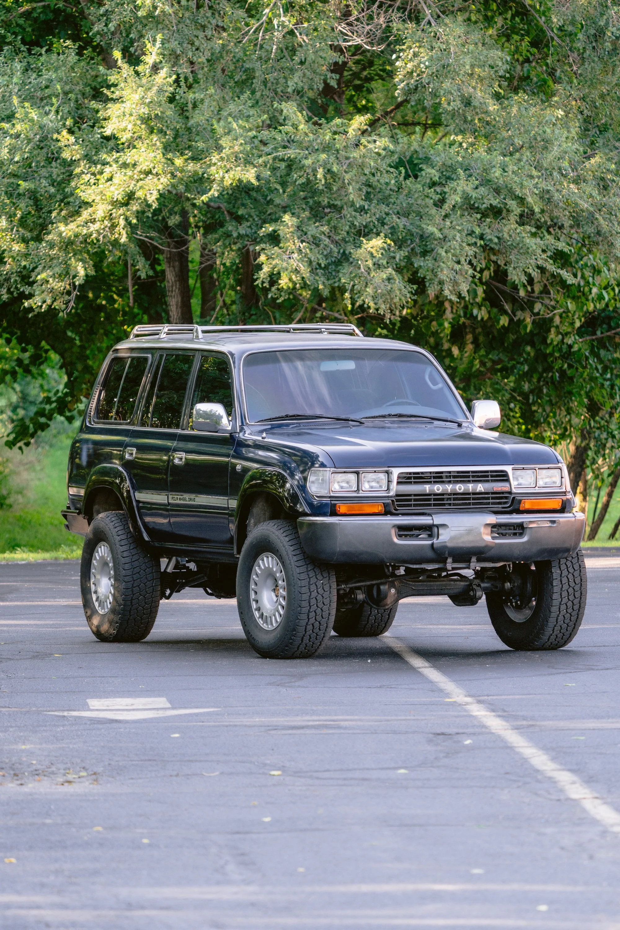 A black Toyota SUV with large tires parked in an empty parking lot, with green trees in the background.