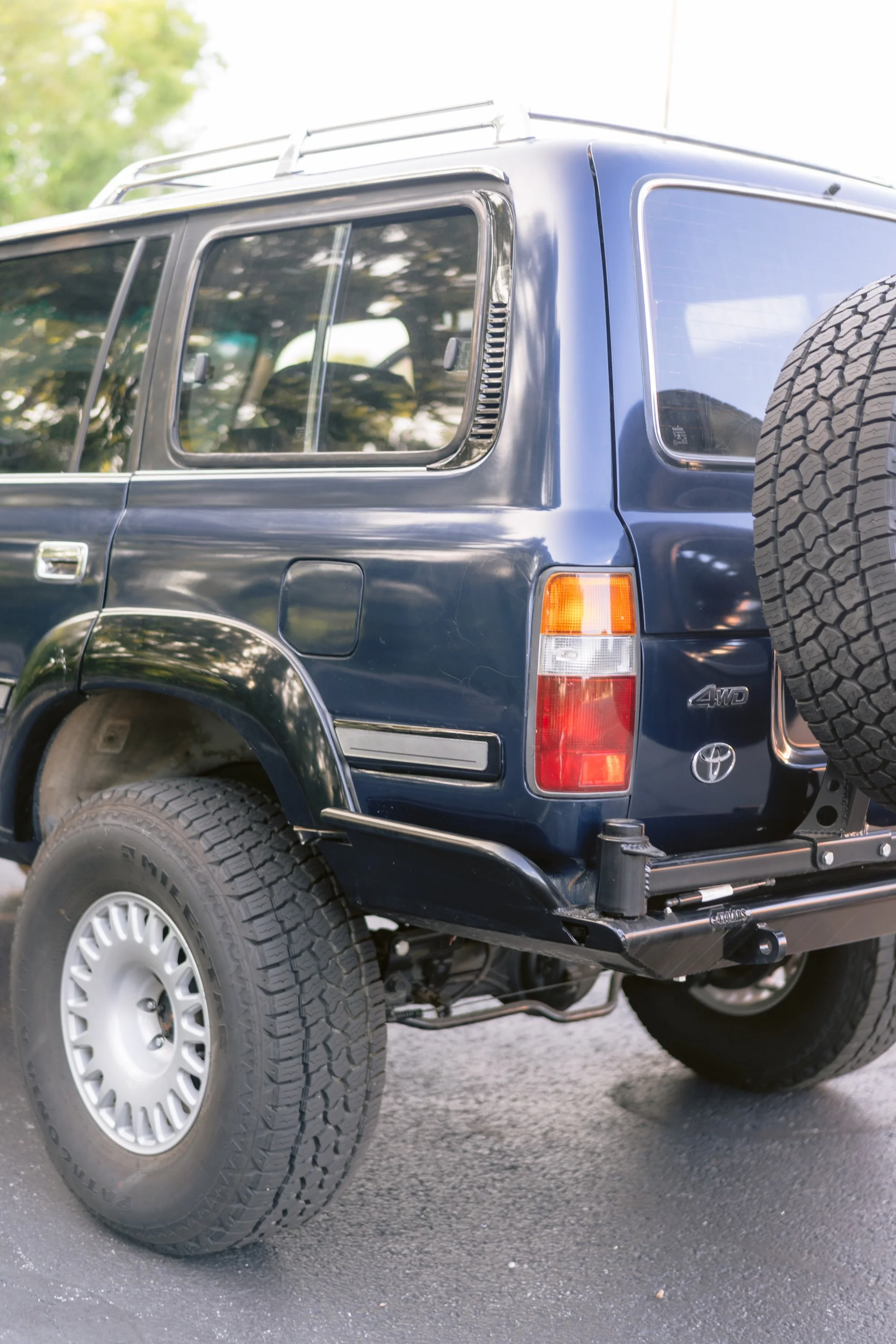 Close-up of the rear and side of a blue Toyota 4WD SUV, showing large off-road tires, a spare tire mounted on the back, and a roof rack. The vehicle is parked on a paved surface with trees and sky reflected in the windows.