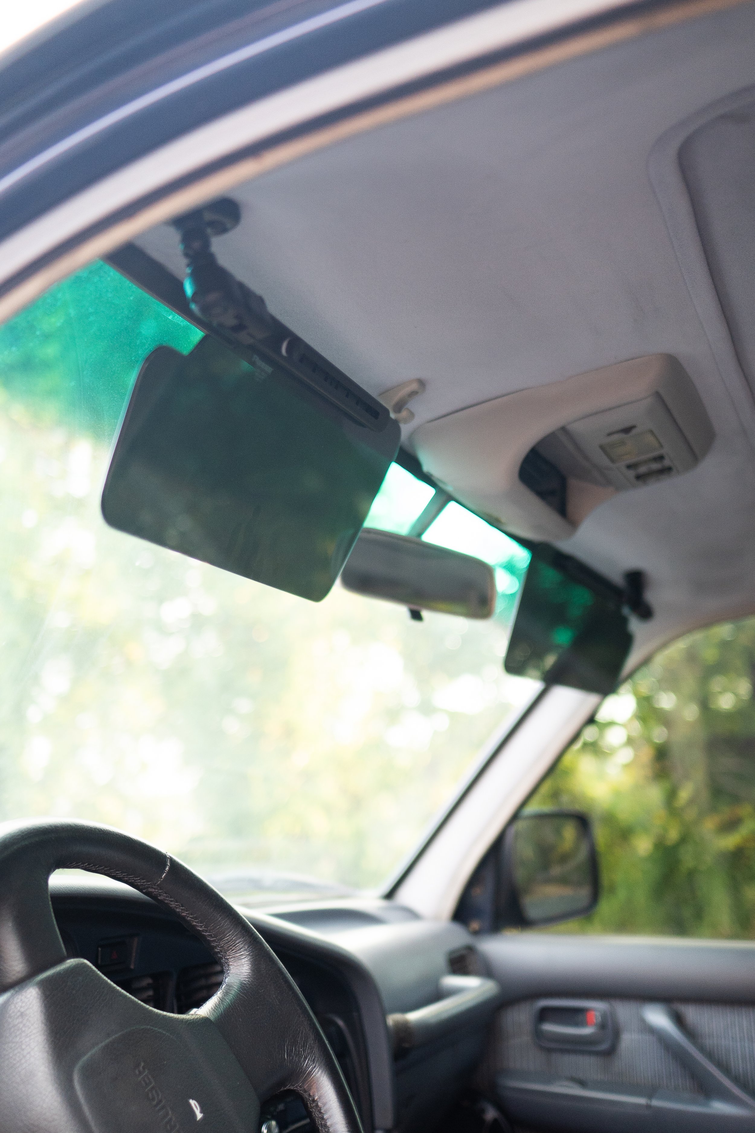 The interior of a vehicle showing a steering wheel, windshield, sun visor, rearview mirror, and part of the dashboard, with green trees visible outside the windshield.