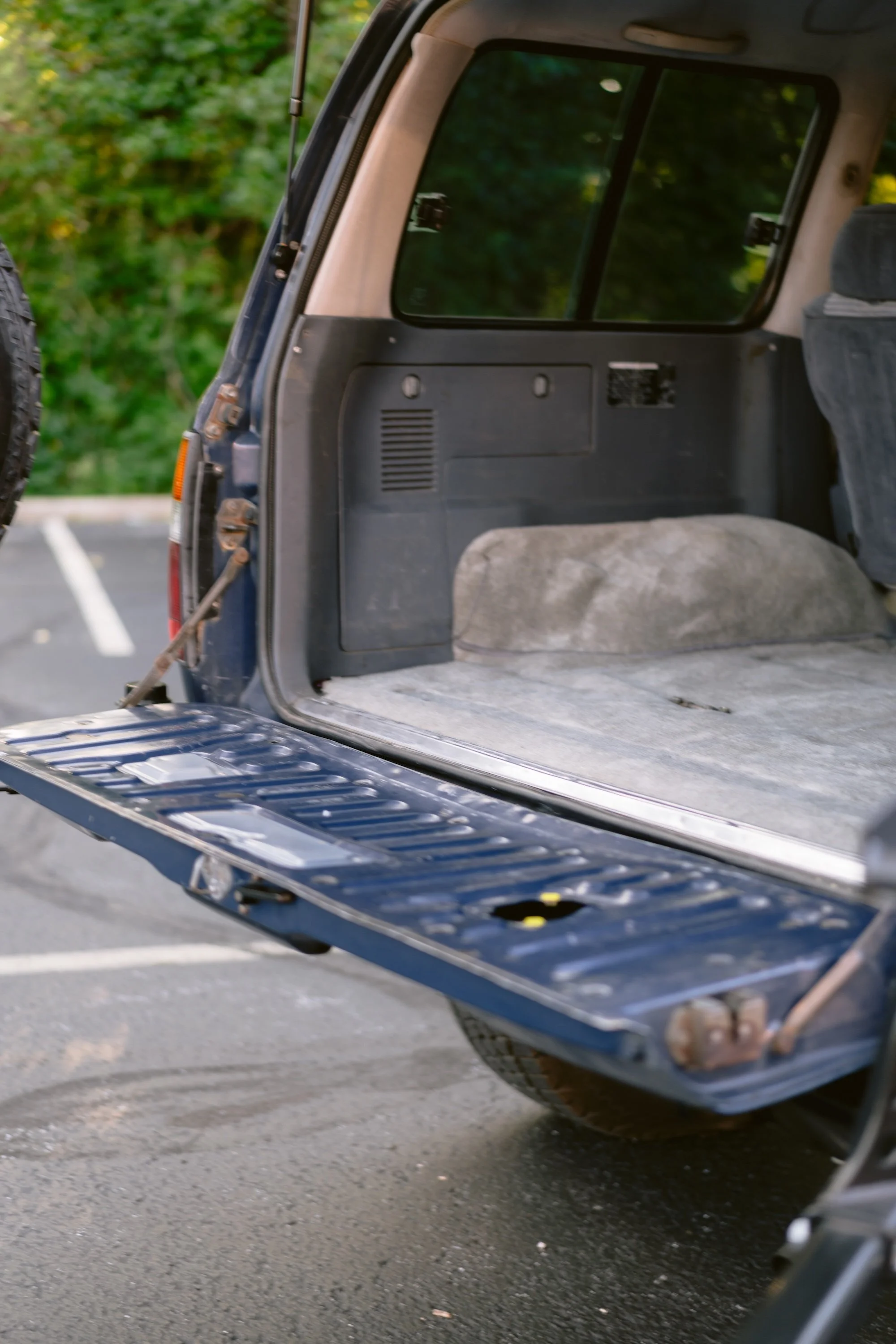 Open rear of a dark blue SUV showing the back seat and trunk area, with a spare tire mounted on the outside left, outside parking lot with trees in the background.