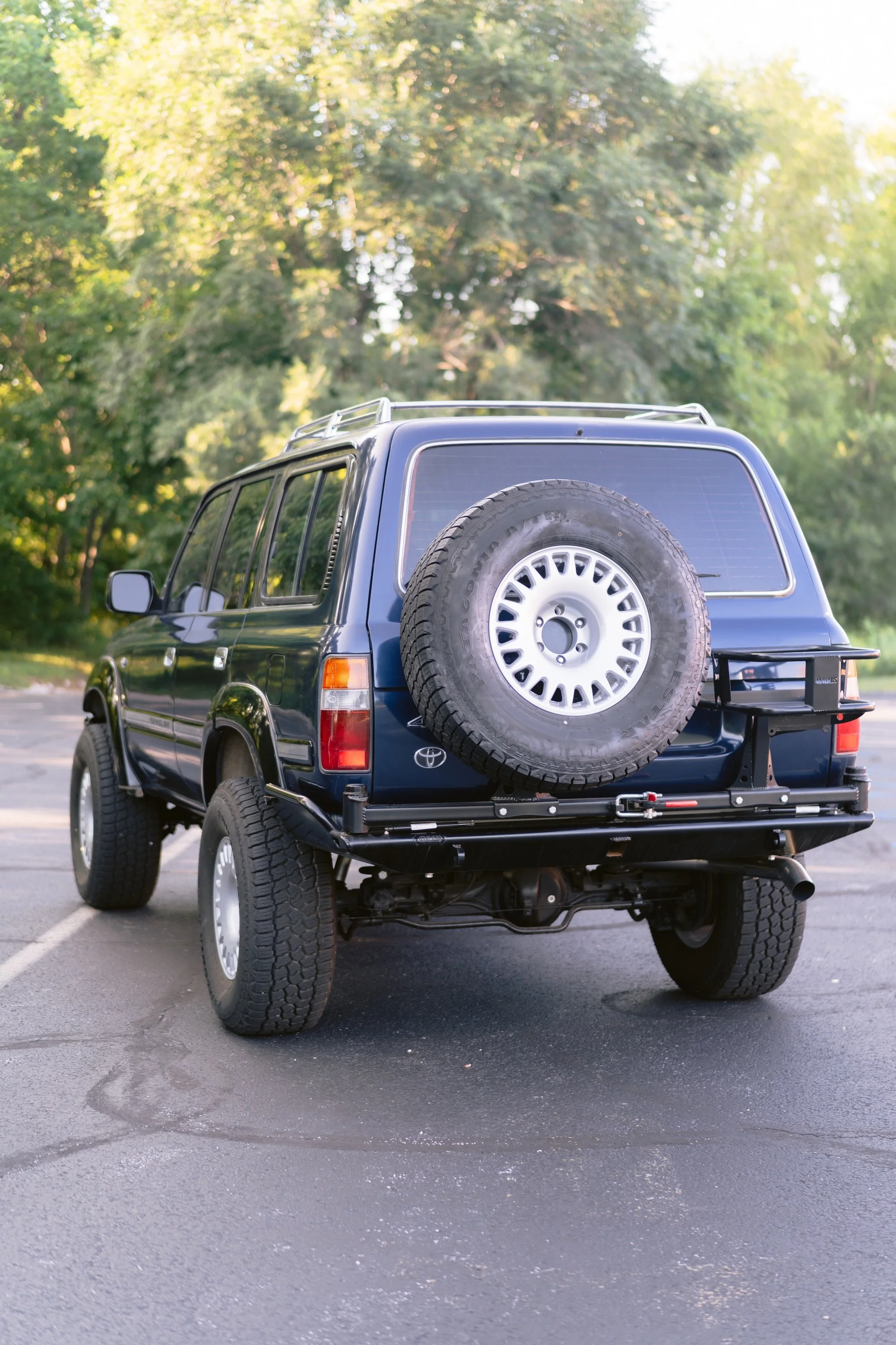 Black SUV with a spare tire mounted on the back, parked on an asphalt surface in front of green trees.