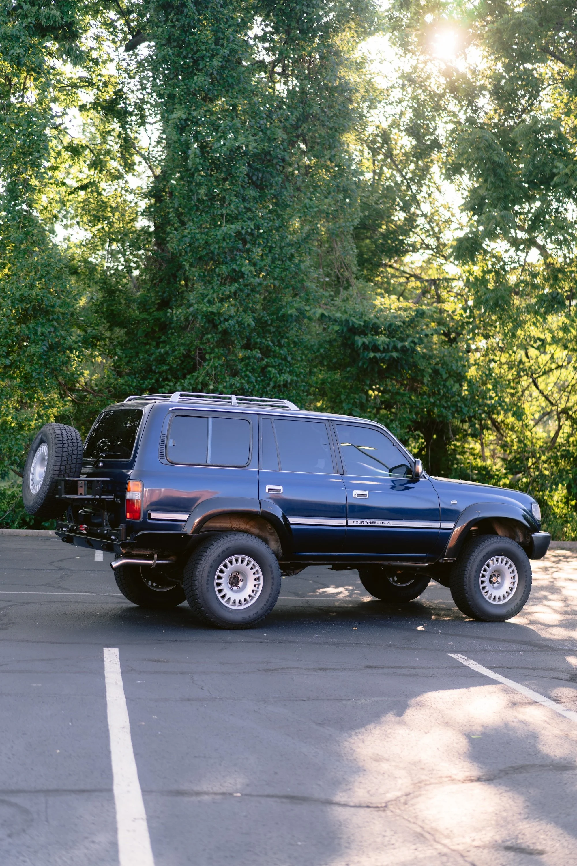 A black SUV with a spare tire mounted on the back, parked in a parking lot with trees and sunlight in the background.