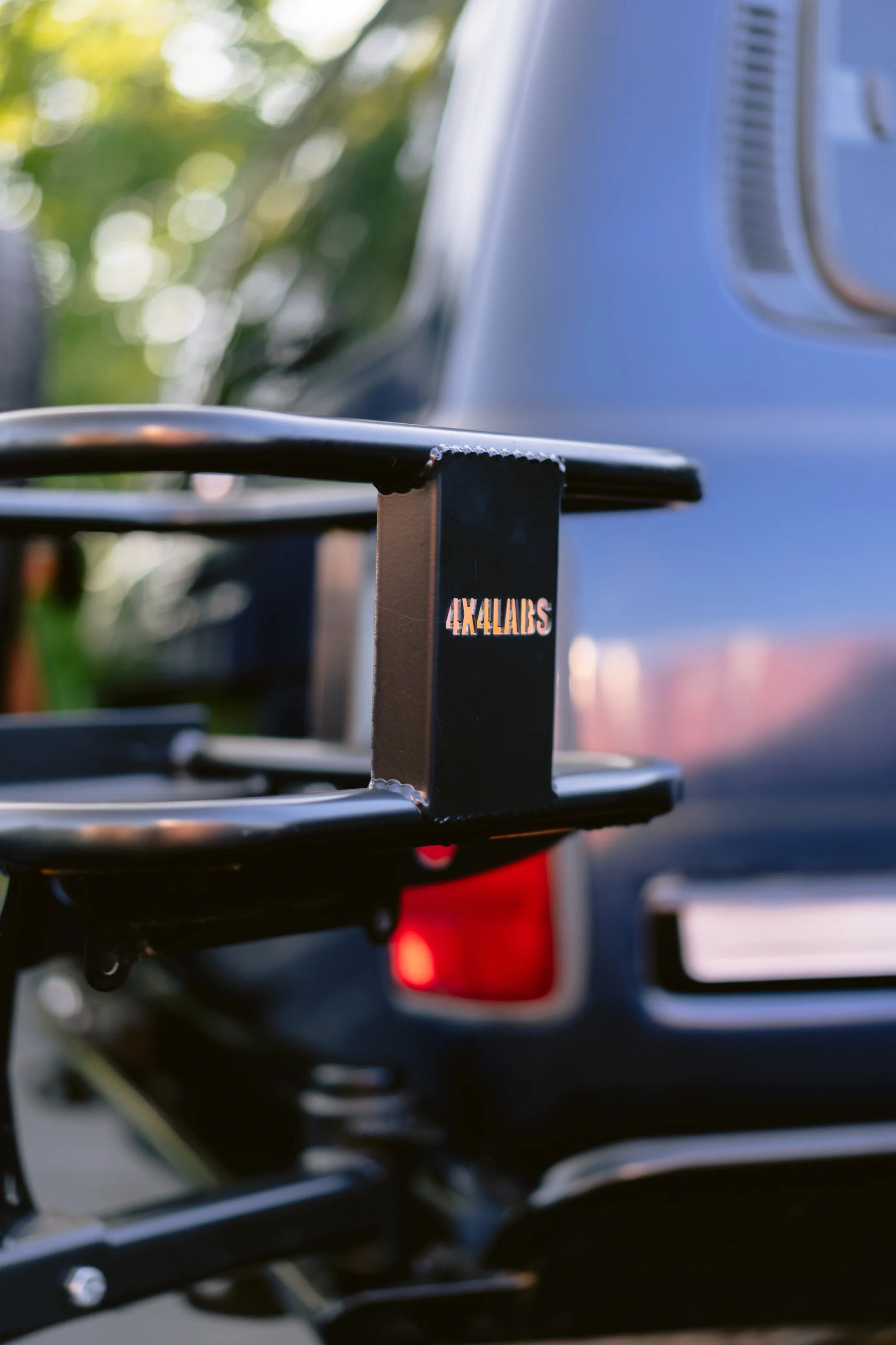 Close-up of a black bicycle rack attached to the back of a vehicle, with September text visible on a small part of the rack, and a blurred background showing a vehicle and greenery.