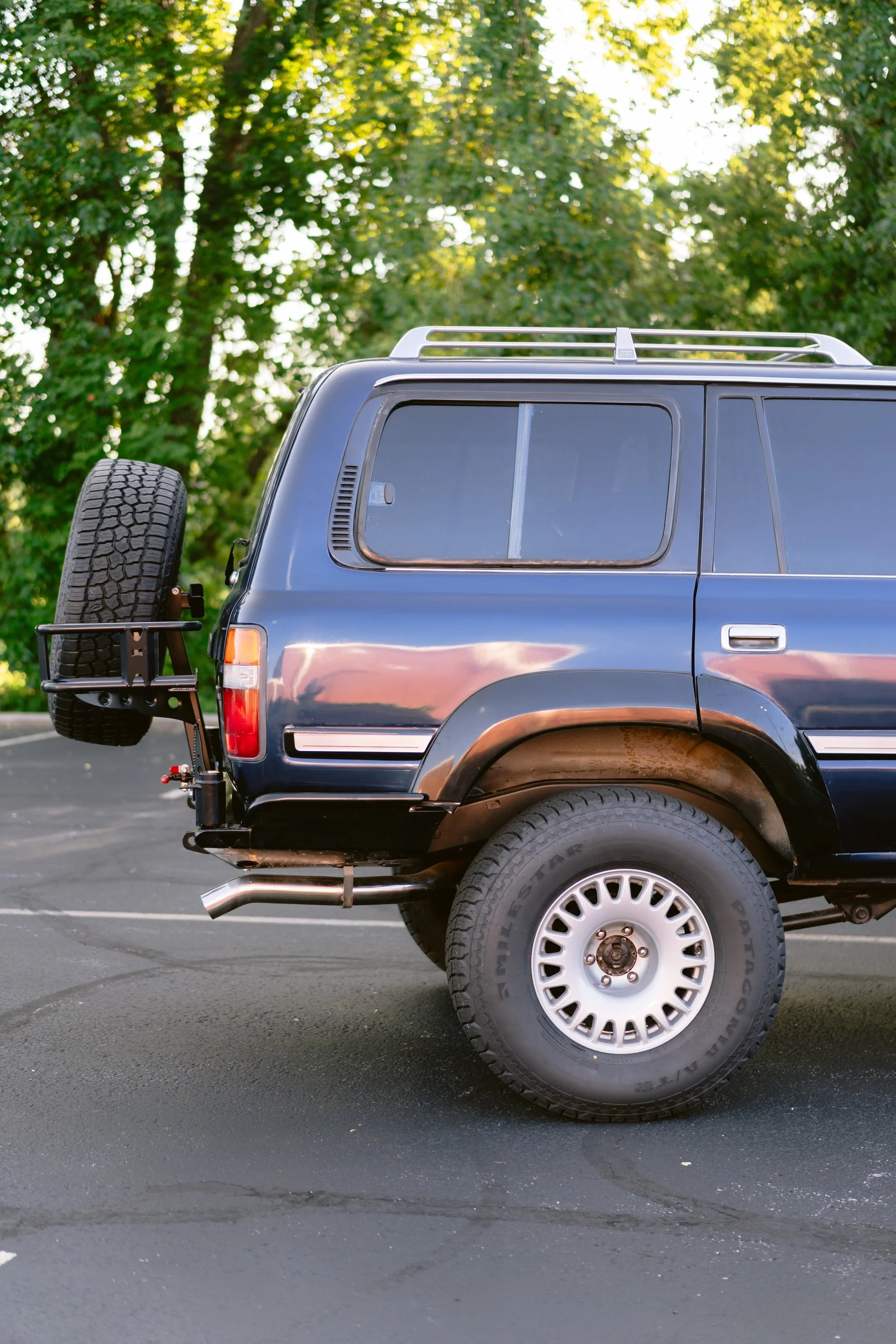 The rear part of a dark-colored SUV parked in a parking lot with green trees in the background, featuring a mounted spare tire and roof rails.