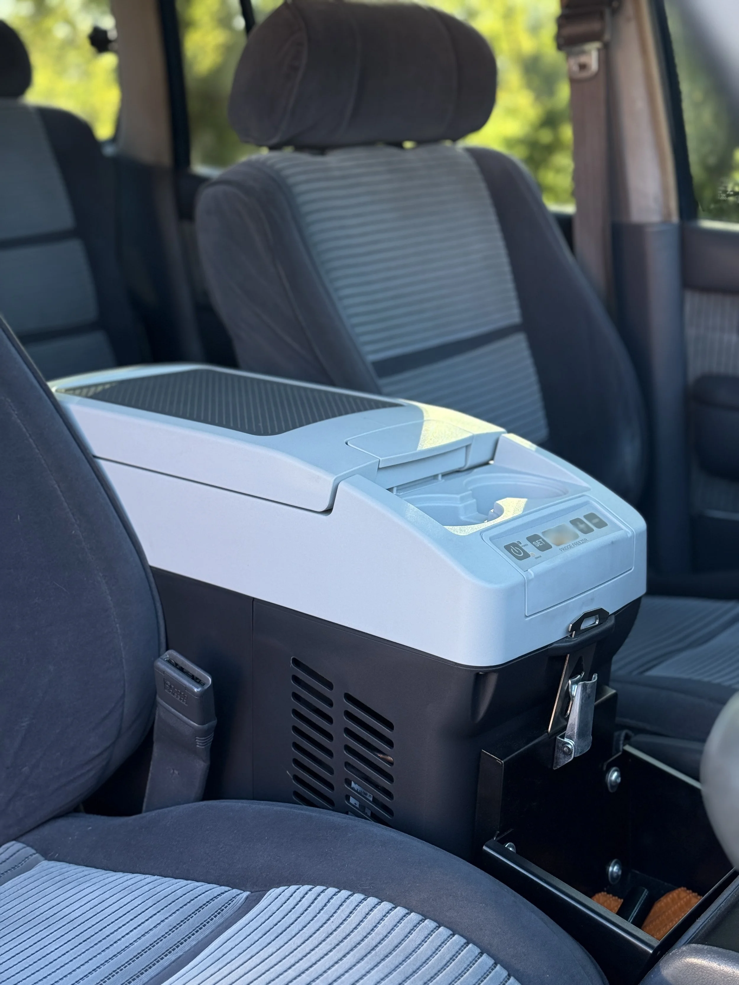 The backseat of a vehicle with a grey fabric seat and a white and grey portable air conditioner or heater placed between the seats.