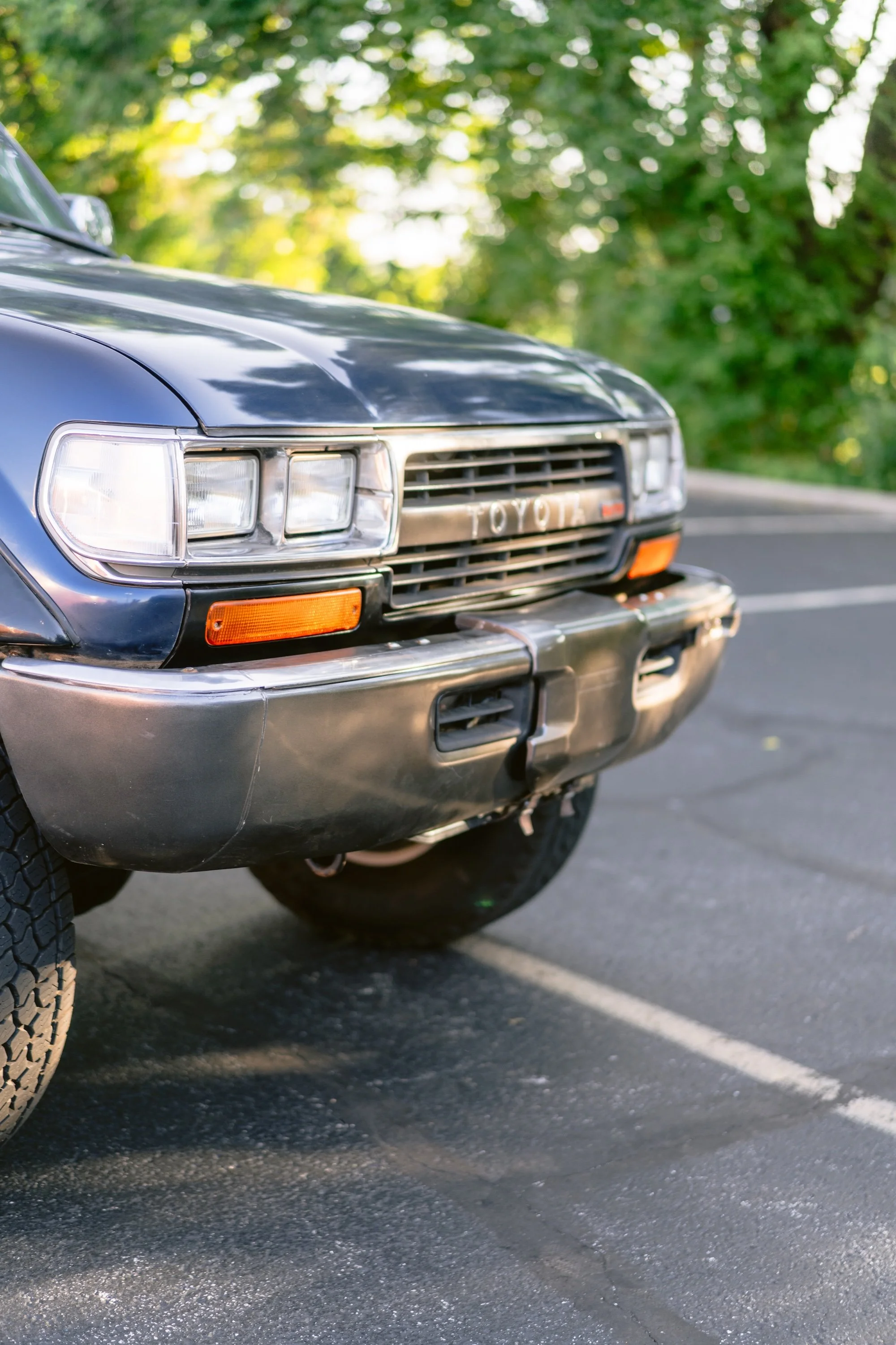 Close-up of the front of a vintage black Toyota SUV parked on a road with a background of green trees and sunlight.