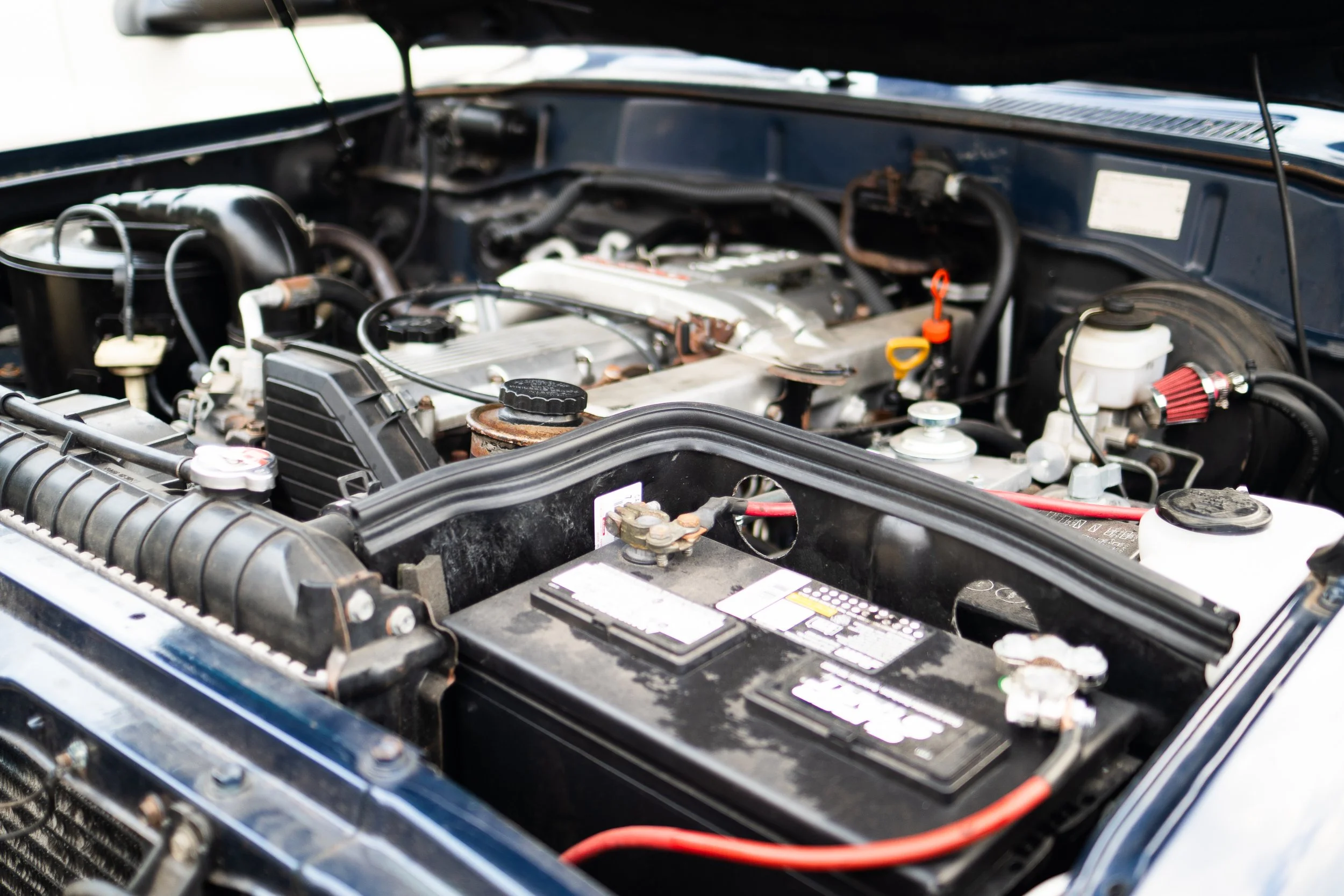 Open car hood revealing engine components, hoses, and battery in an automobile engine bay.
