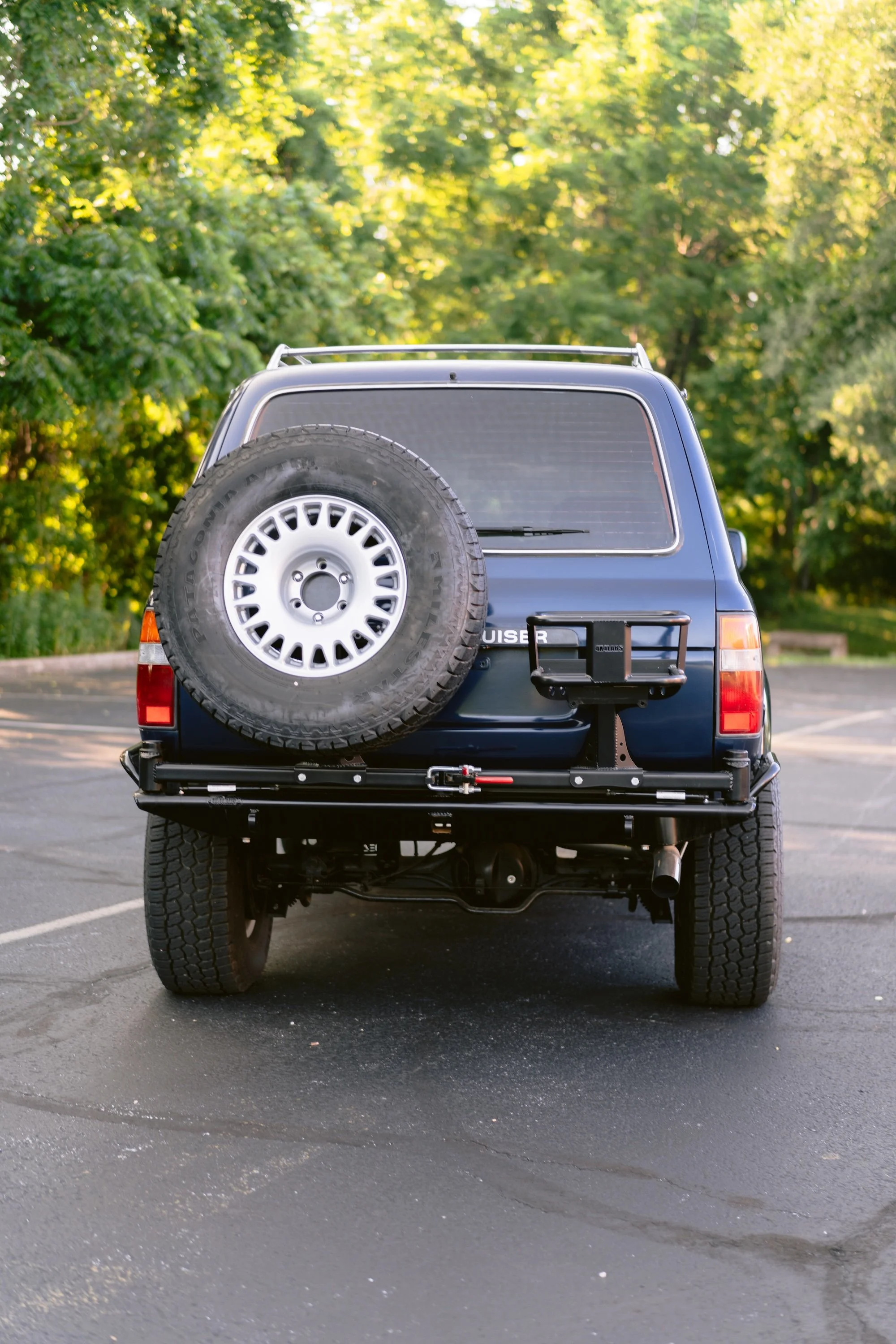 The back of a blue SUV with a spare tire mounted on the rear, parked in an outdoor parking lot with trees and greenery in the background.