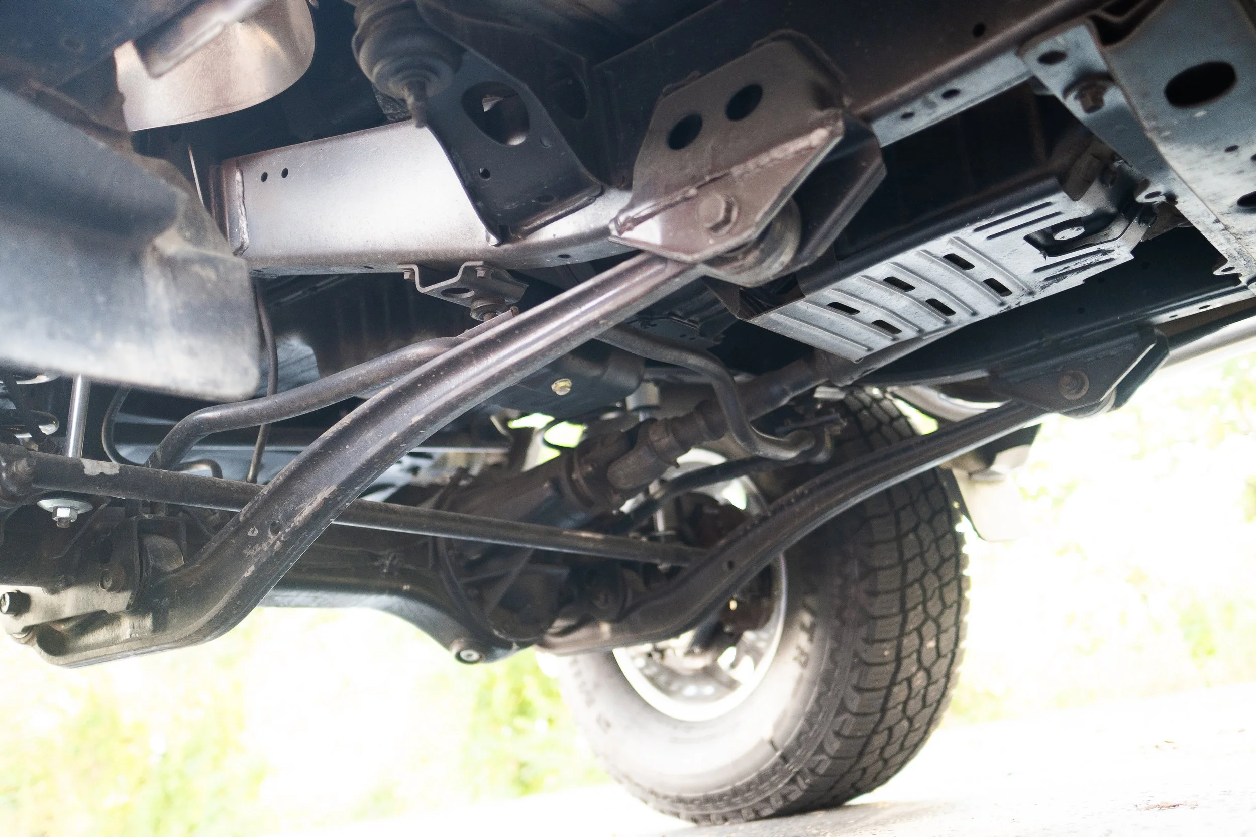 Underneath view of a vehicle's chassis showing suspension components, metal frame, engine parts, and a tire in the background.