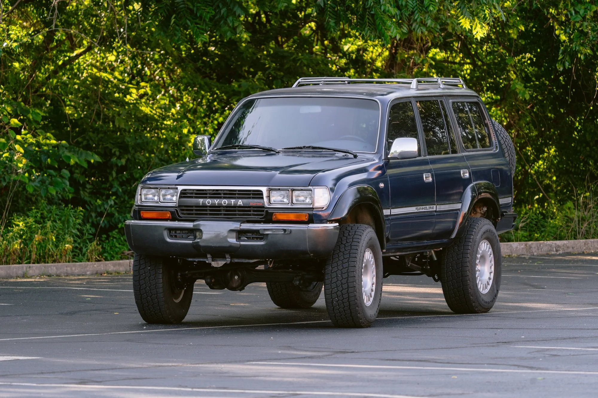A black Toyota 4x4 SUV with a roof rack, parked in a lot with a background of green trees and bushes.