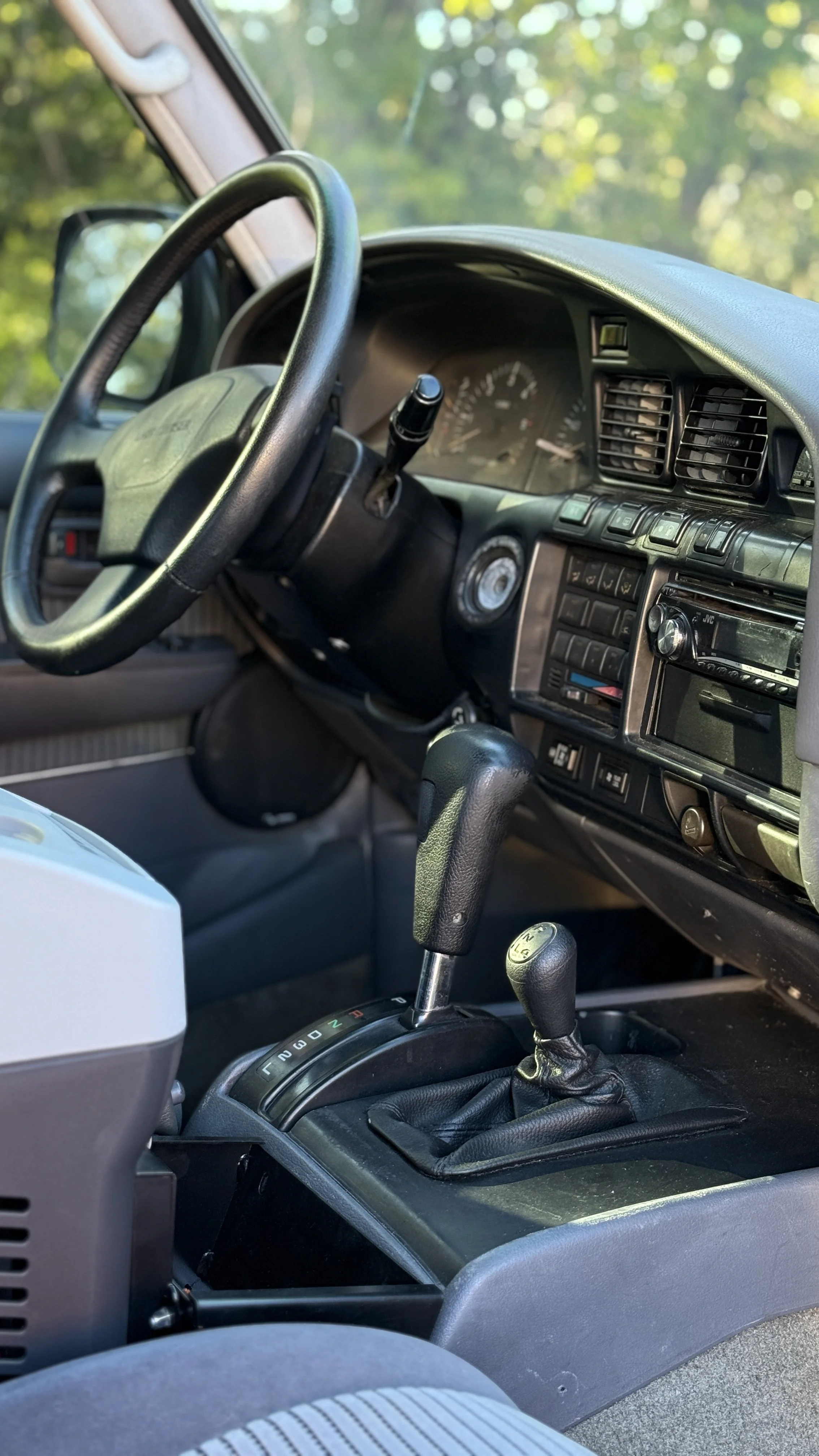 The interior of a vehicle showing the steering wheel, dashboard, gear shift, and some controls, with a blurred view of green trees outside the window.