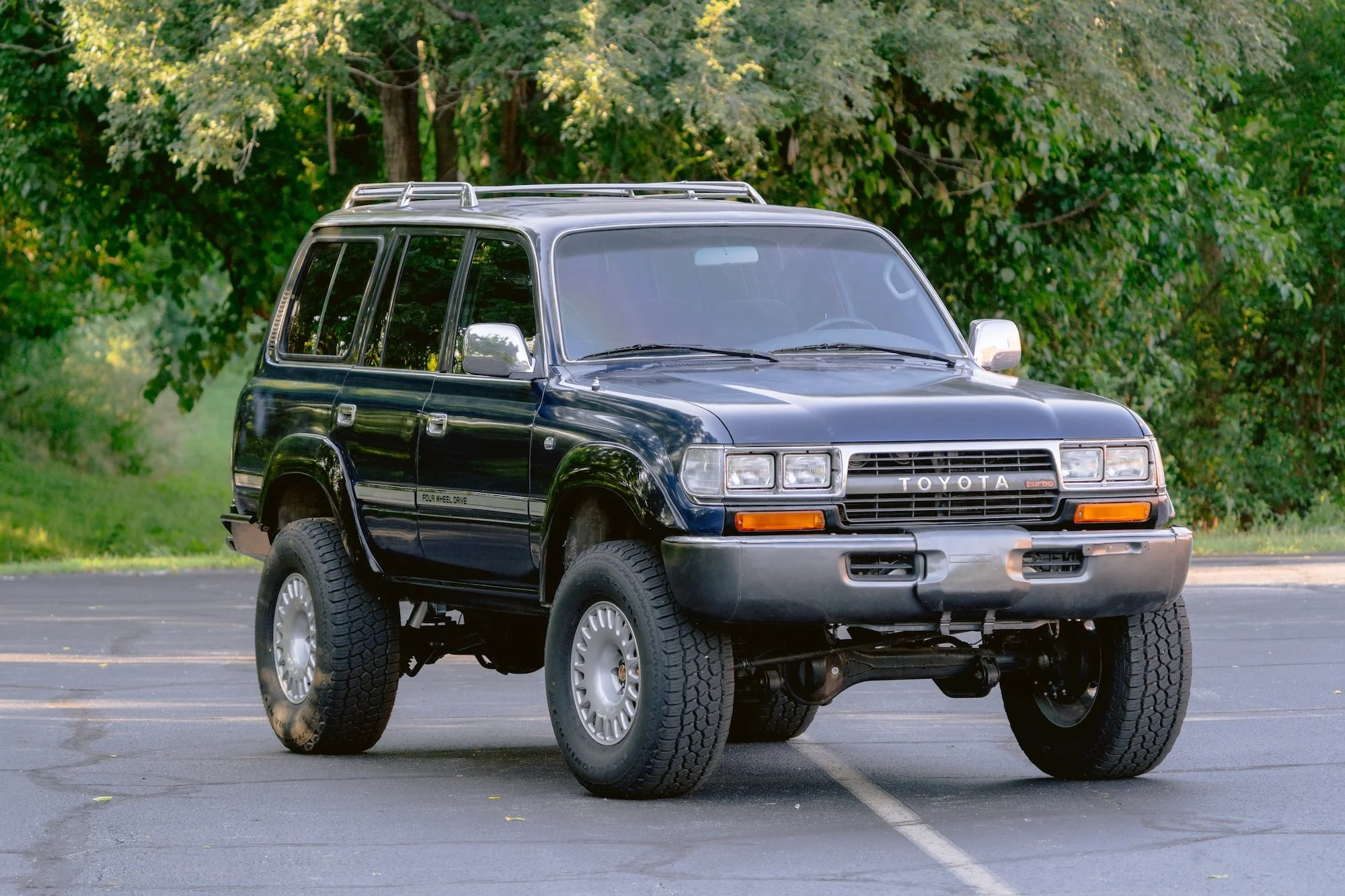 A black Toyota SUV with the front wheels and part of the body lifted in the air, parked on an asphalt lot with trees and greenery in the background.