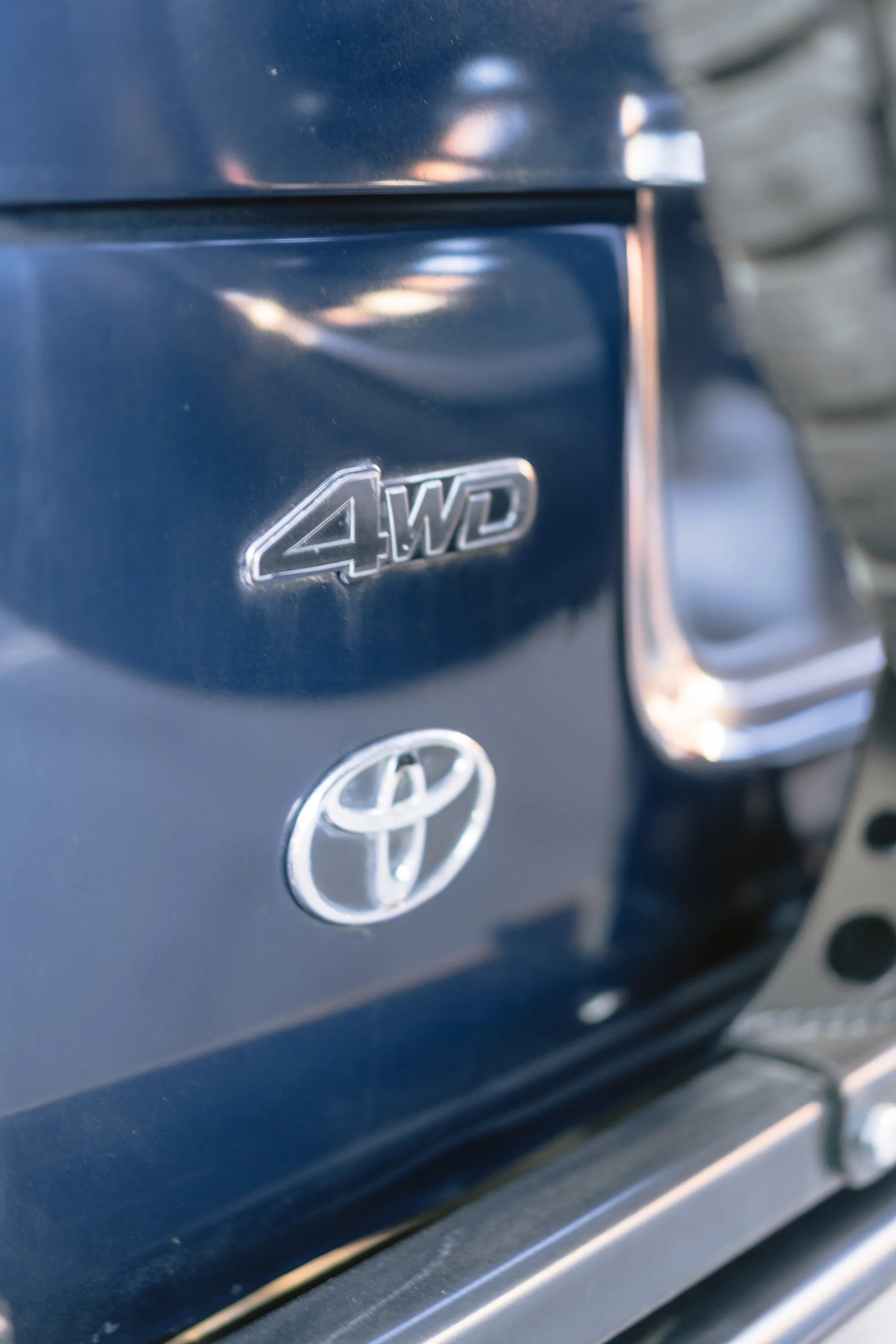 Close-up of the back of a vehicle showing a silver 4WD badge and a silver Toyota logo, with a blurred background.