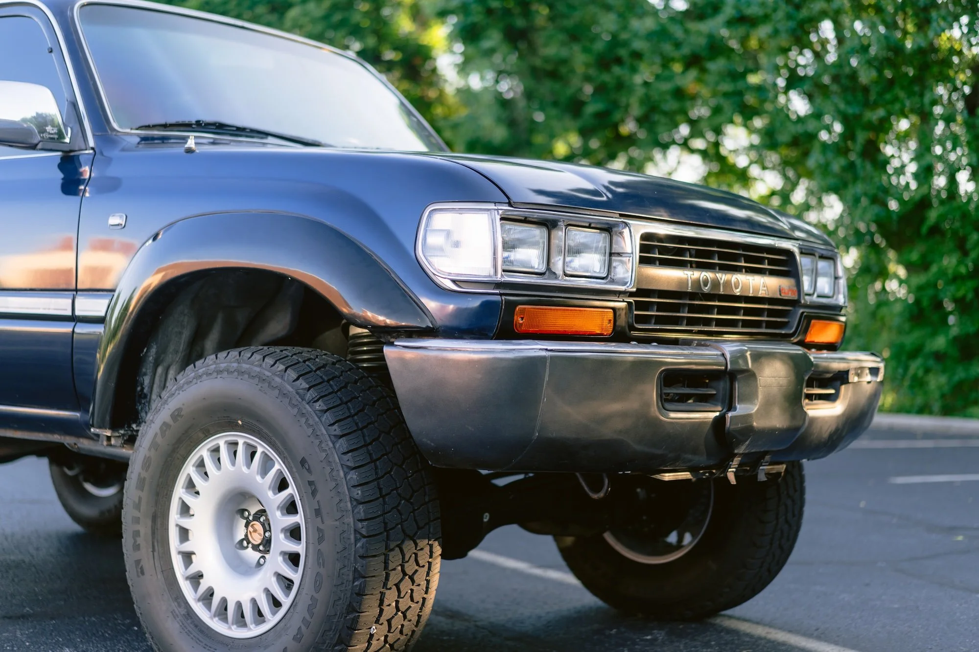 Close-up of a vintage blue Toyota pickup truck parked outdoors with green trees in the background.