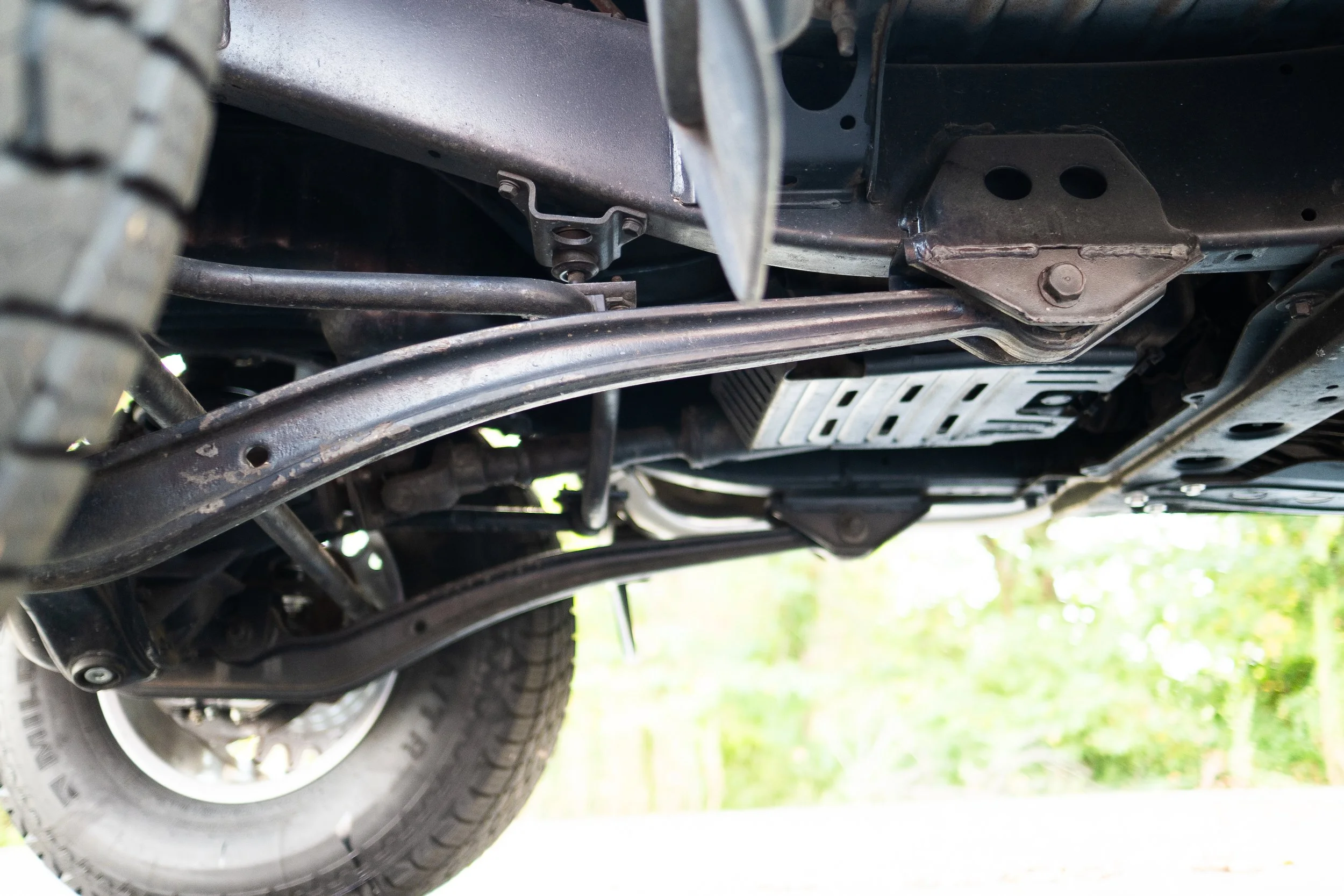 Underneath view of a vehicle showing suspension components, metal frame parts, and part of the tire with a blurred background of grass.