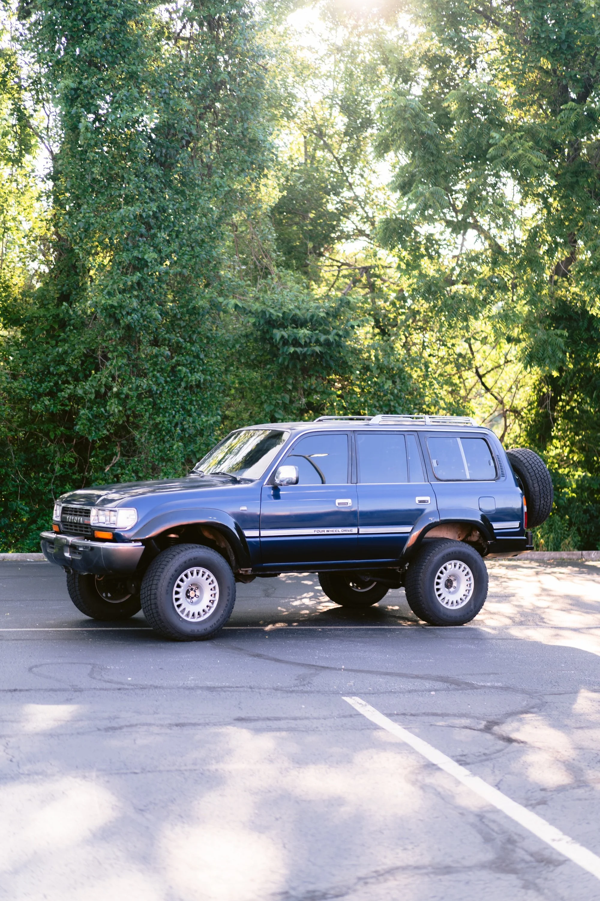 A blue Toyota SUV with large off-road tires parked in a parking lot surrounded by trees with bright green leaves.