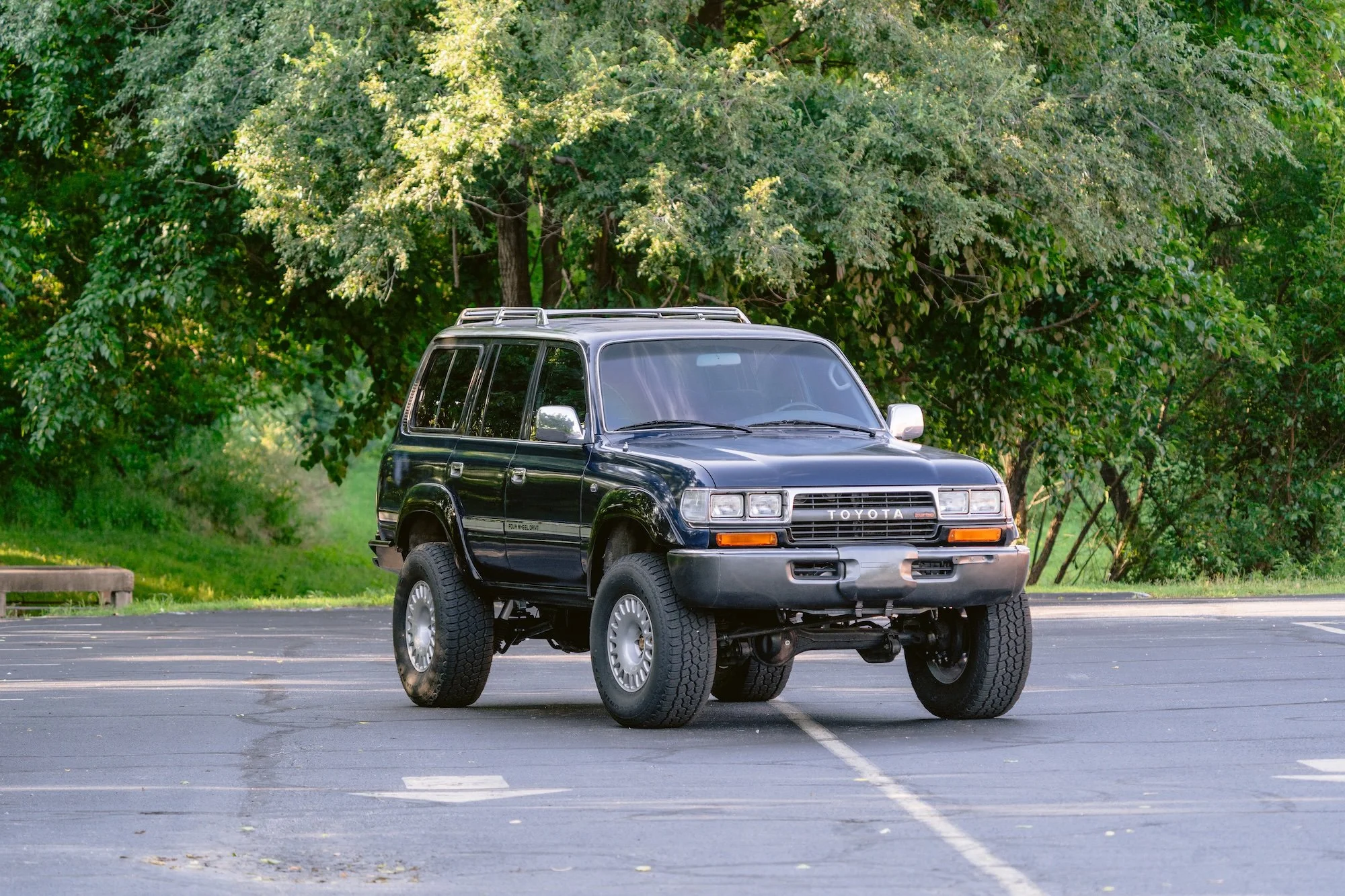 A black Toyota SUV with large tires is parked in an empty parking lot with green trees in the background.