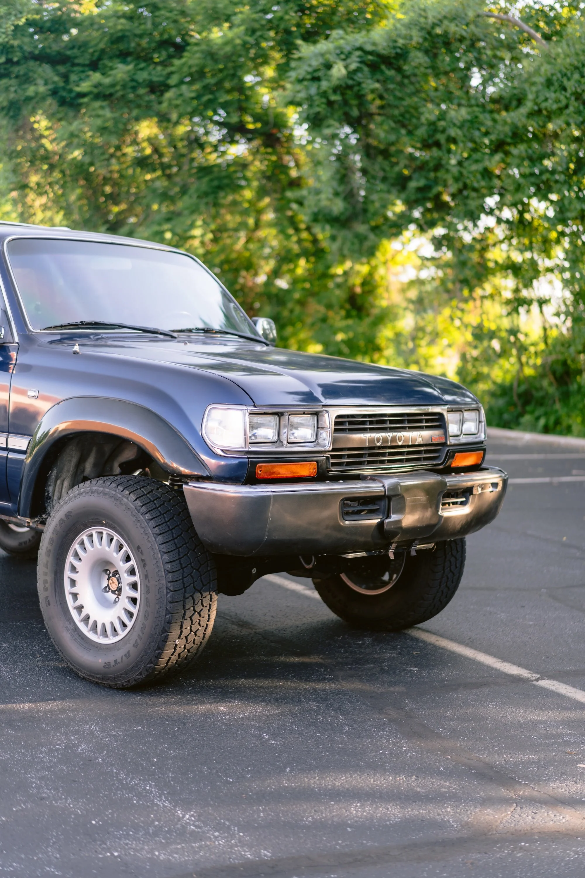 A black vintage Toyota SUV parked in an outdoor lot with a background of green trees and sunlight filtering through the leaves.
