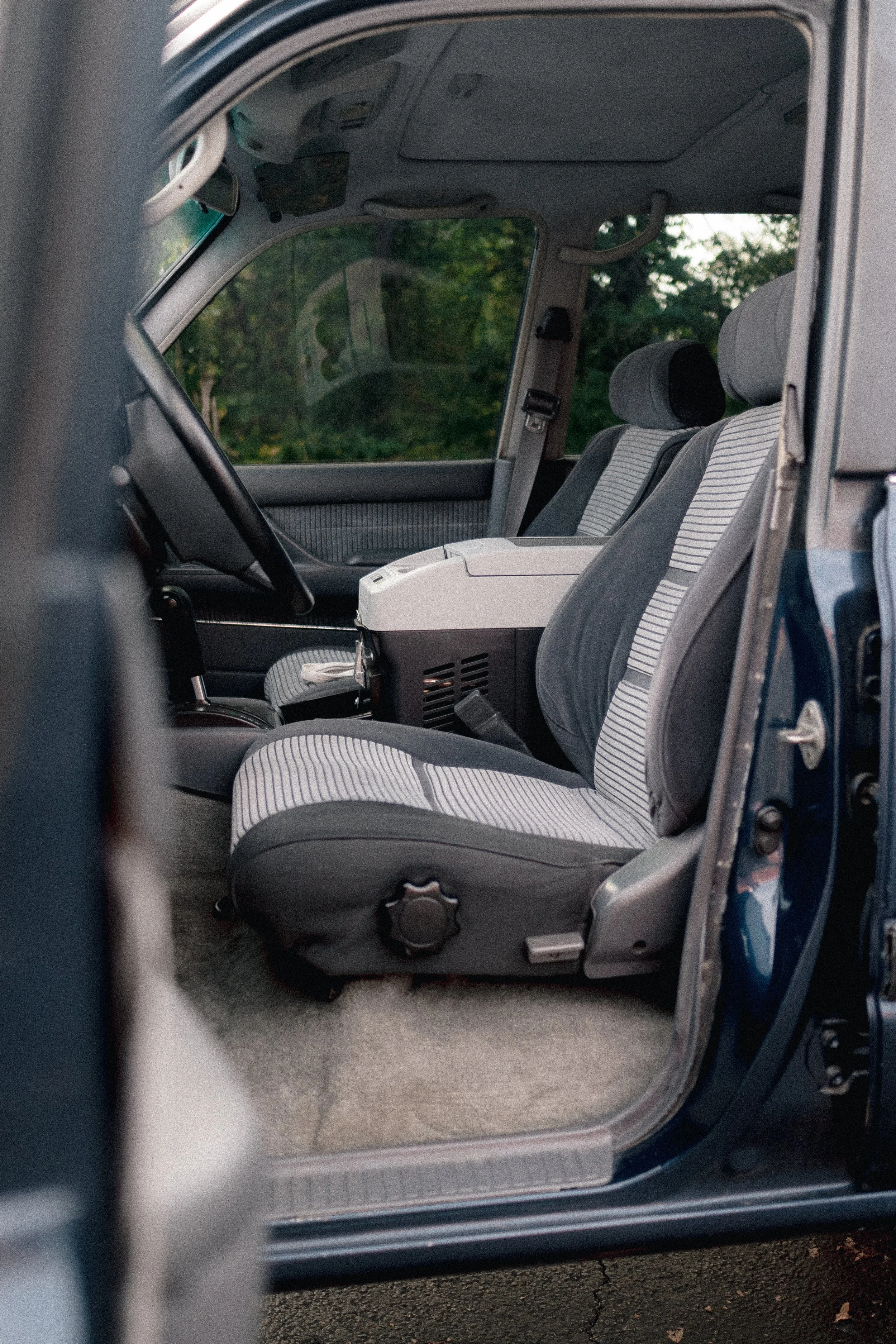 Inside view of a car's passenger side showing two front seats with gray and black striped upholstery, a central console, and a view through the window of trees outside.