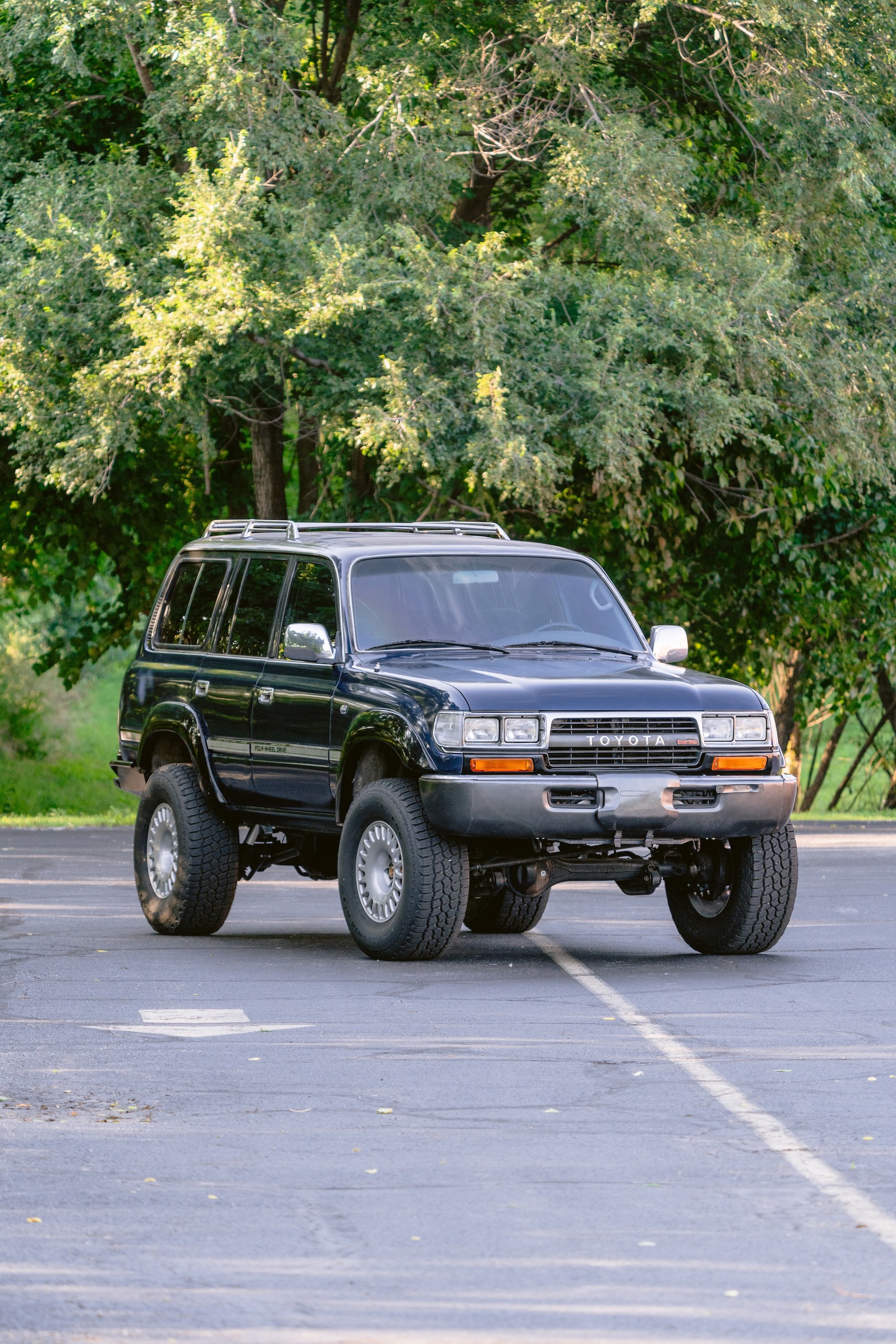 A black Toyota SUV is parked in an empty parking lot with yellow lines on the asphalt. Behind it, there are green trees and bushes.