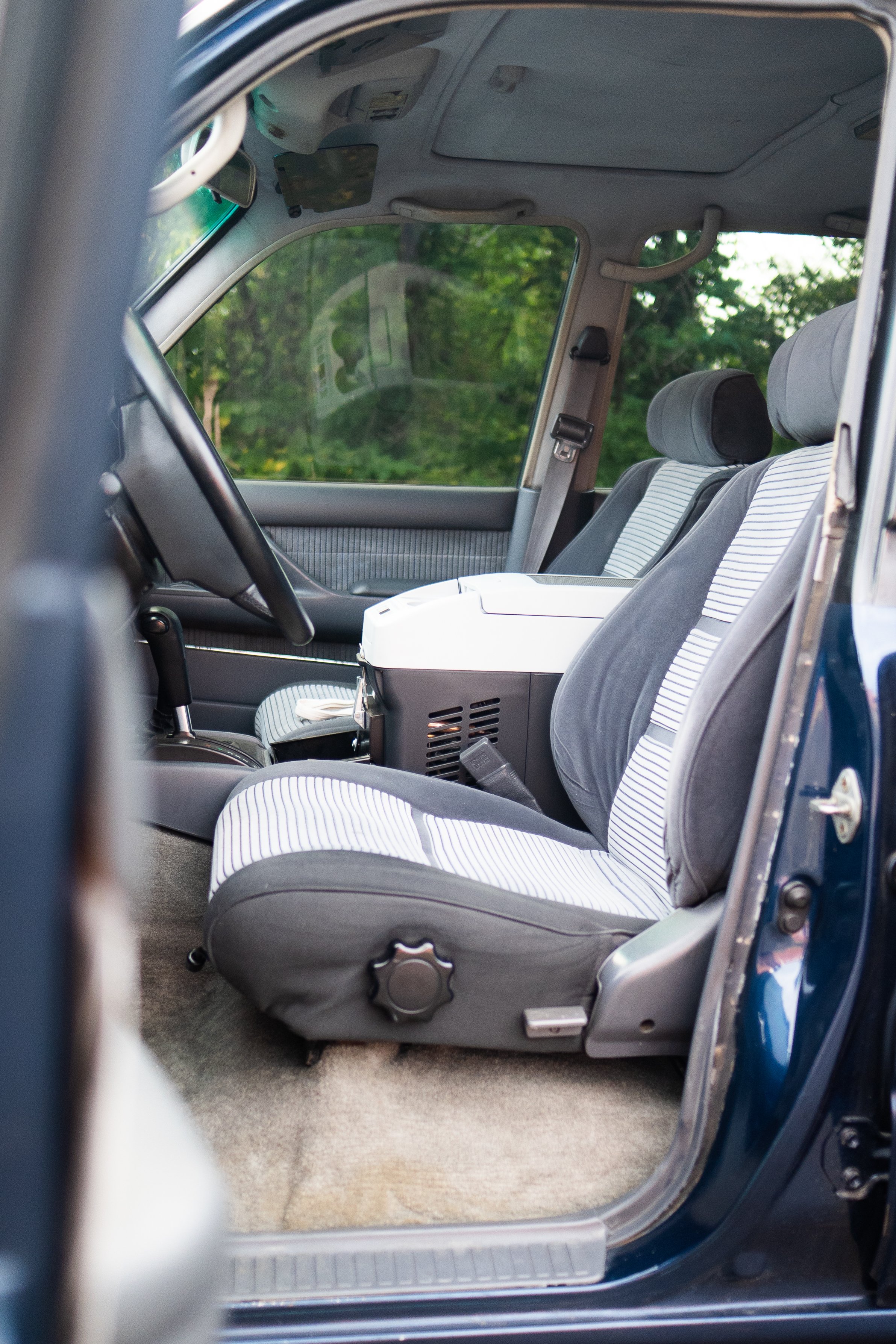 The interior of a car showing the front seats, dashboard, and part of the steering wheel, with a view through the open driver side door to the lush green trees outside.
