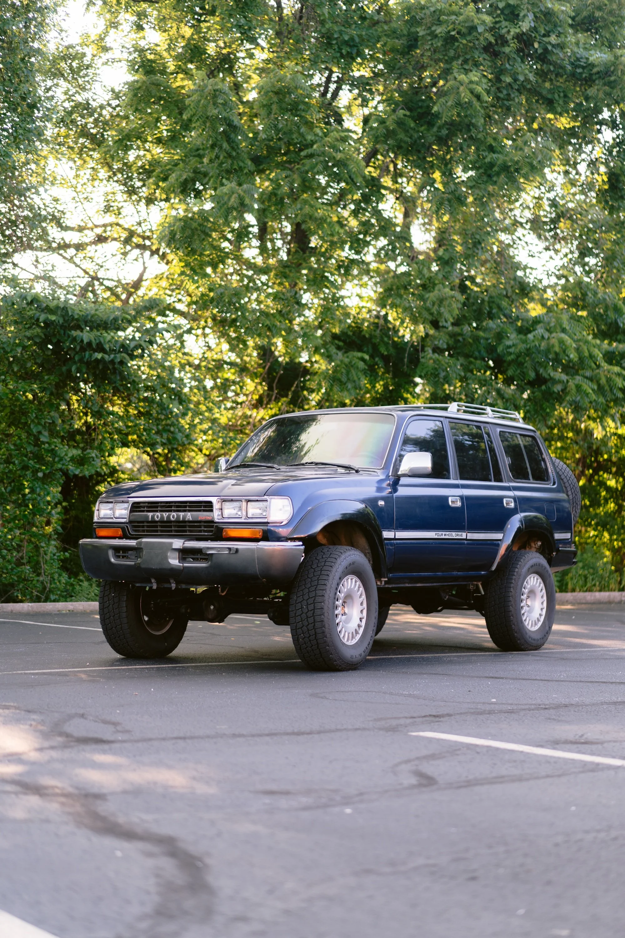 A dark blue SUV with large tires parked in a nearly empty parking lot surrounded by trees with lush green leaves.