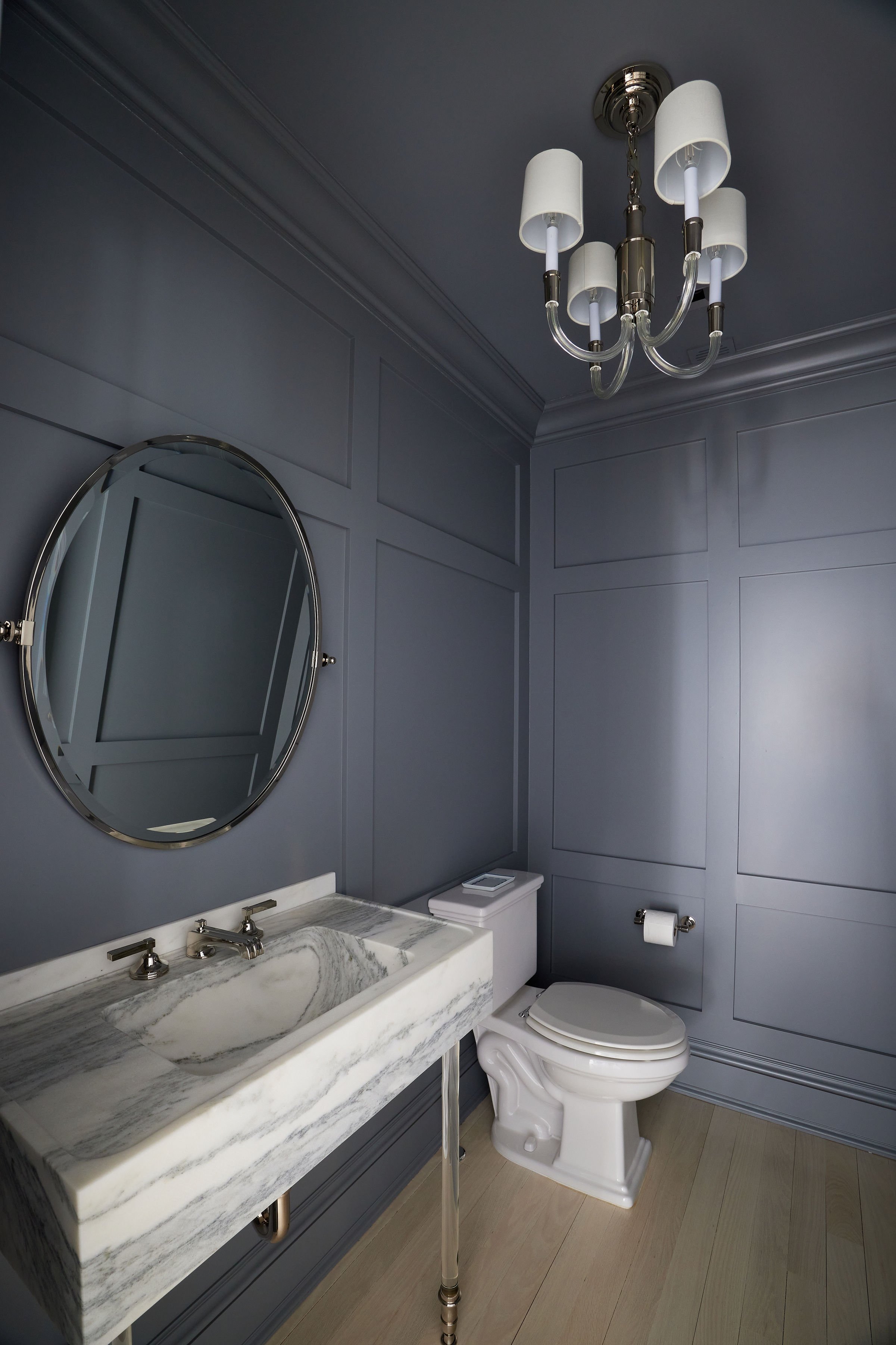 A bathroom with gray paneled walls, a marble countertop with a vessel sink, a round mirror, a white toilet, and a chandelier with six white lampshades hanging from the ceiling.