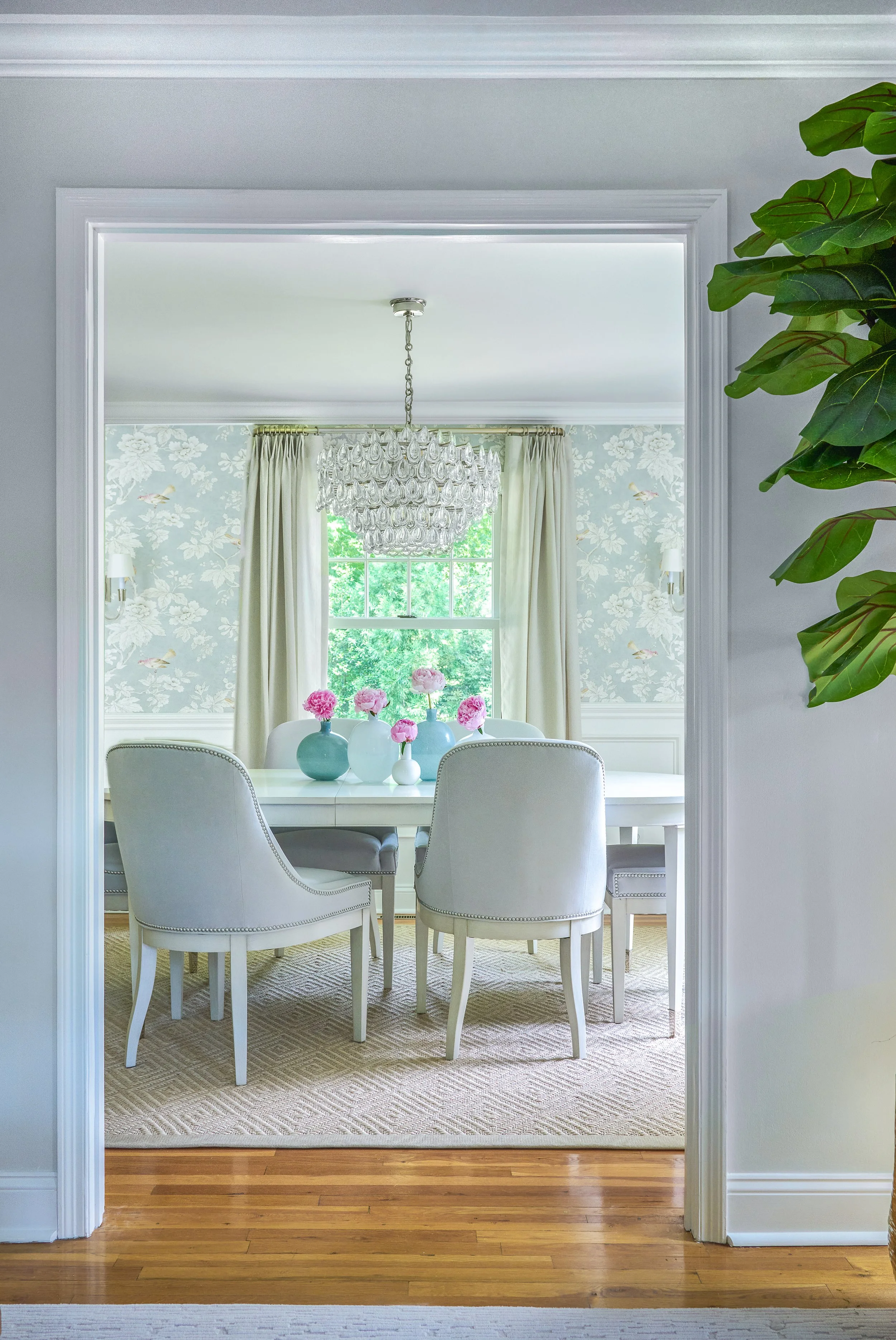 A dining room with a white table, six white upholstered chairs with nailhead trim, pink flowers in vases, a chandelier, large window with curtains, and floral wallpaper.