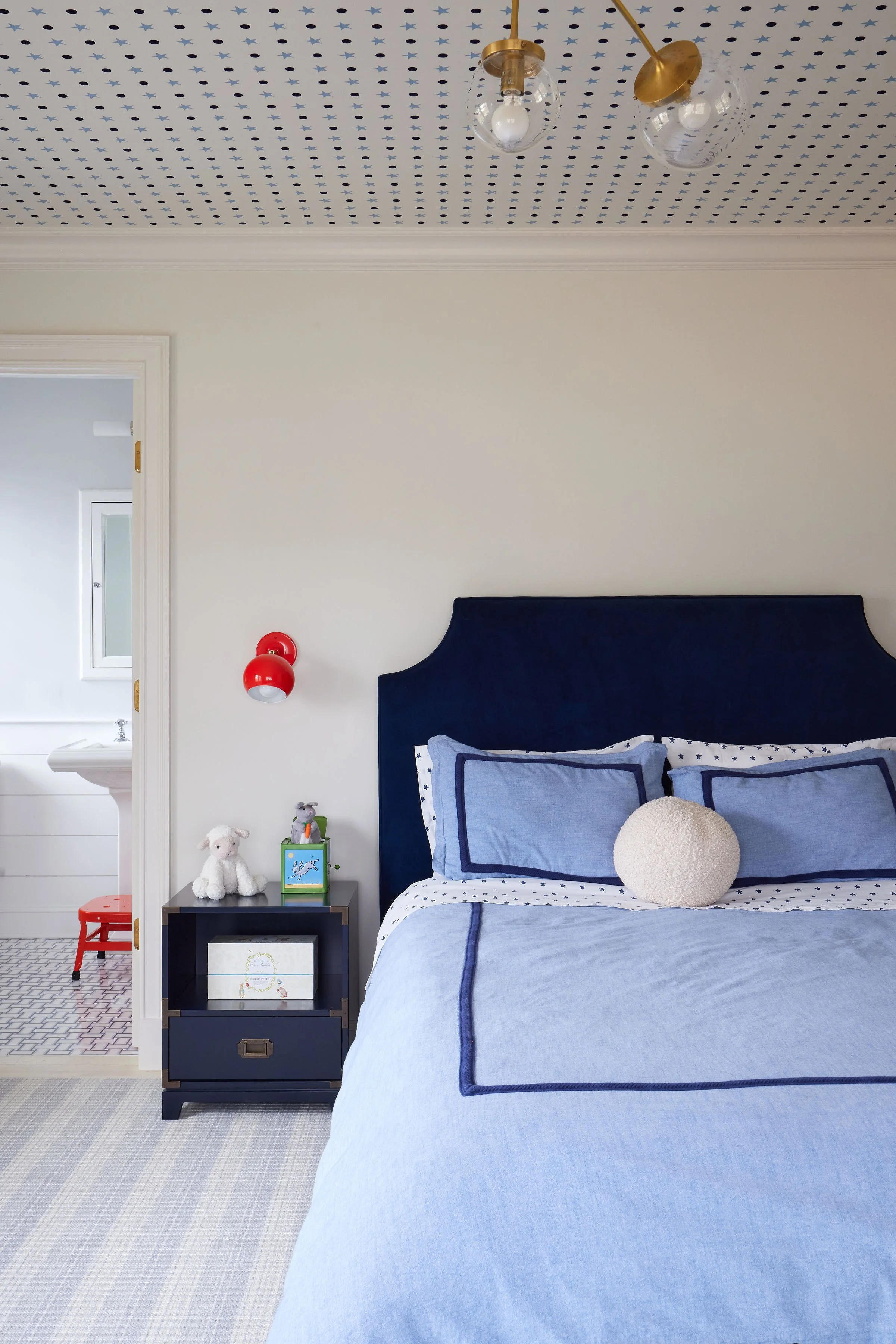 A bedroom with light blue bedding, a dark blue headboard, a beige round pillow, and a corner nightstand with stuffed animals, a small green box, and a framed picture. The wall is beige, and there's a white doorway leading to a bathroom with a sink. The ceiling has a blue star-patterned wallpaper, and there are two red wall-mounted lights and a modern ceiling light fixture.