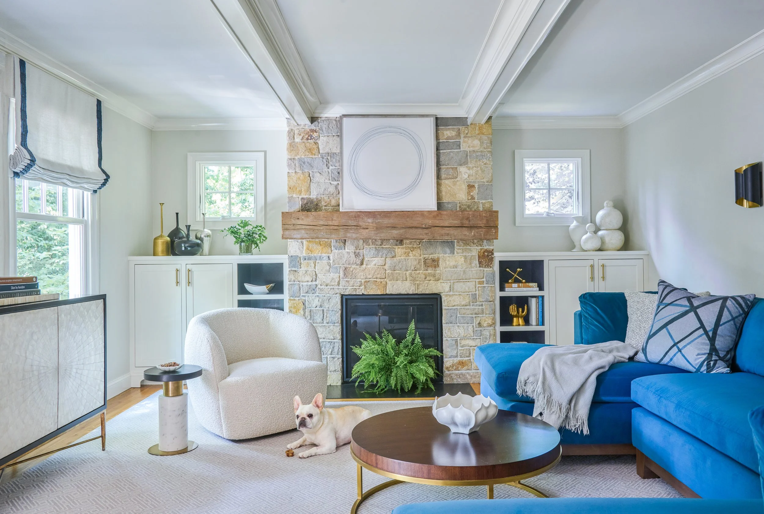 Living room with a blue sofa, white armchair, and a French bulldog on the carpet. There is a stone and brick fireplace with a wooden mantel, decorated with framed artwork and vases. Large windows with white frames and roman shades let in natural light. A round wooden coffee table with a white decorative bowl is in front of the sofa, and a white sideboard with books and decor is against the wall.