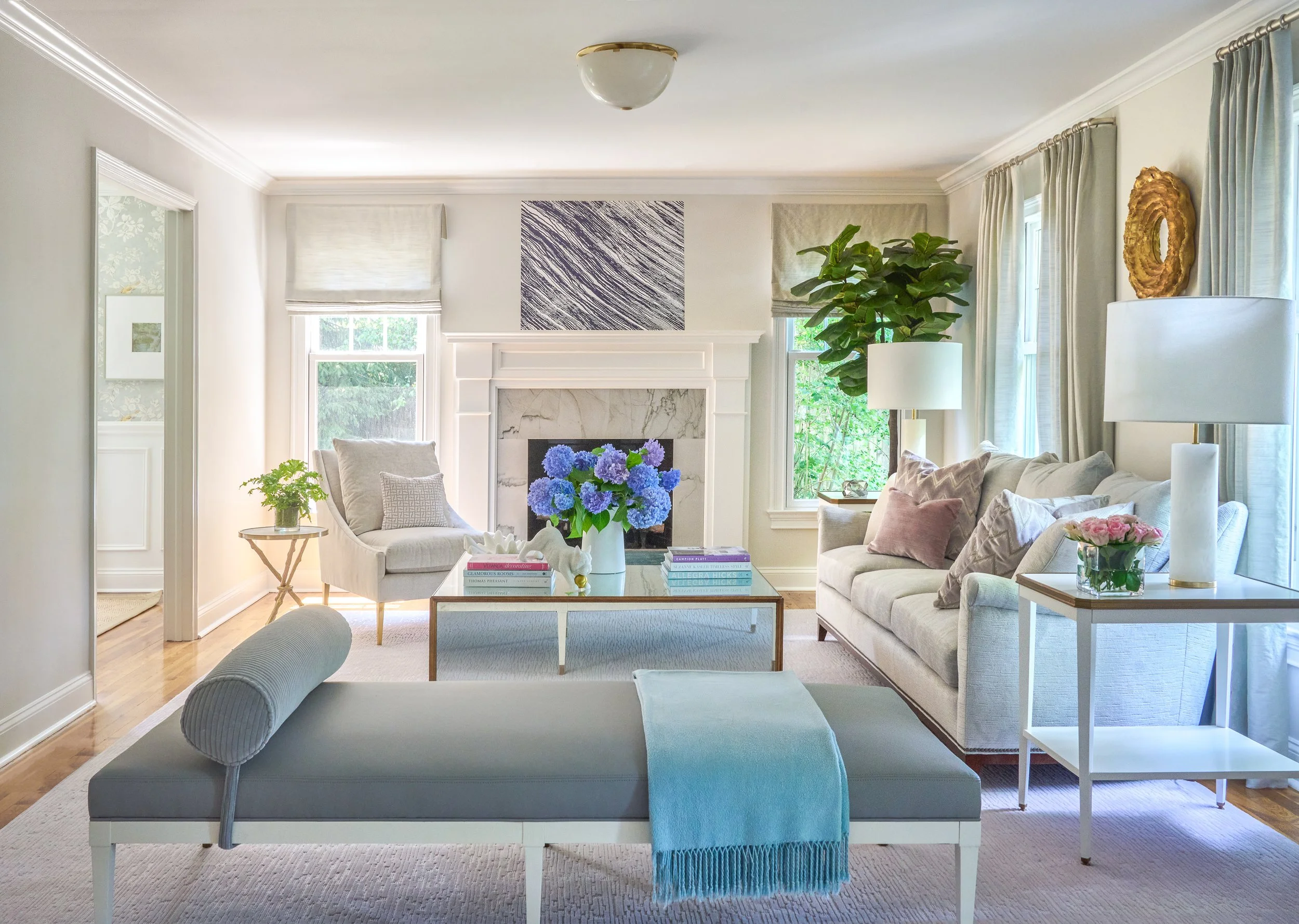 A cozy living room with a white fireplace, blue hydrangea flowers on a coffee table, and beige and pastel-colored furniture. There are large windows with white curtains and decorative wall art.