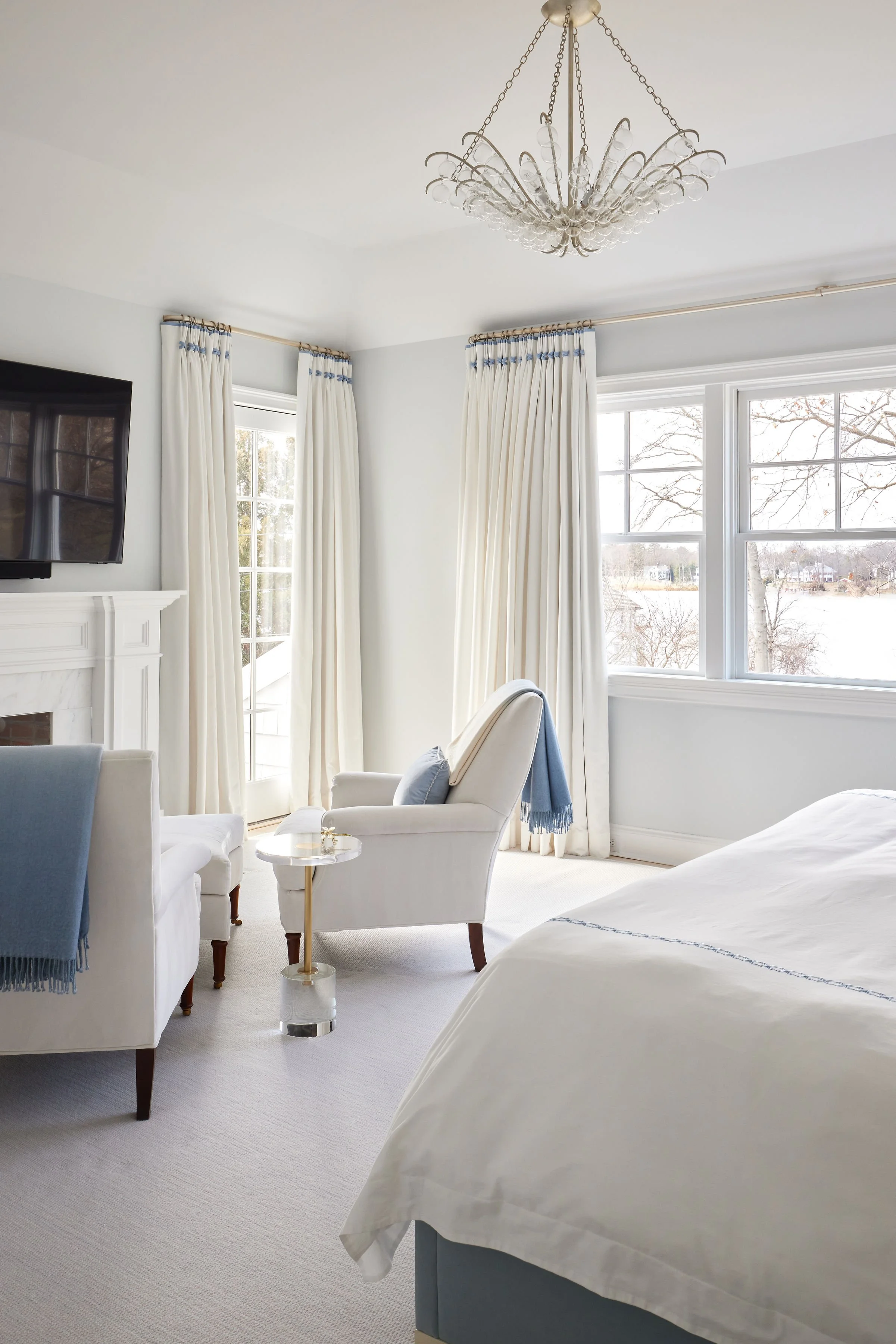 A bright master bedroom sitting area with white walls, large windows with white curtains, a chandelier overhead, a white armchair with a blue pillow, a side table, and part of a bed visible in the foreground.