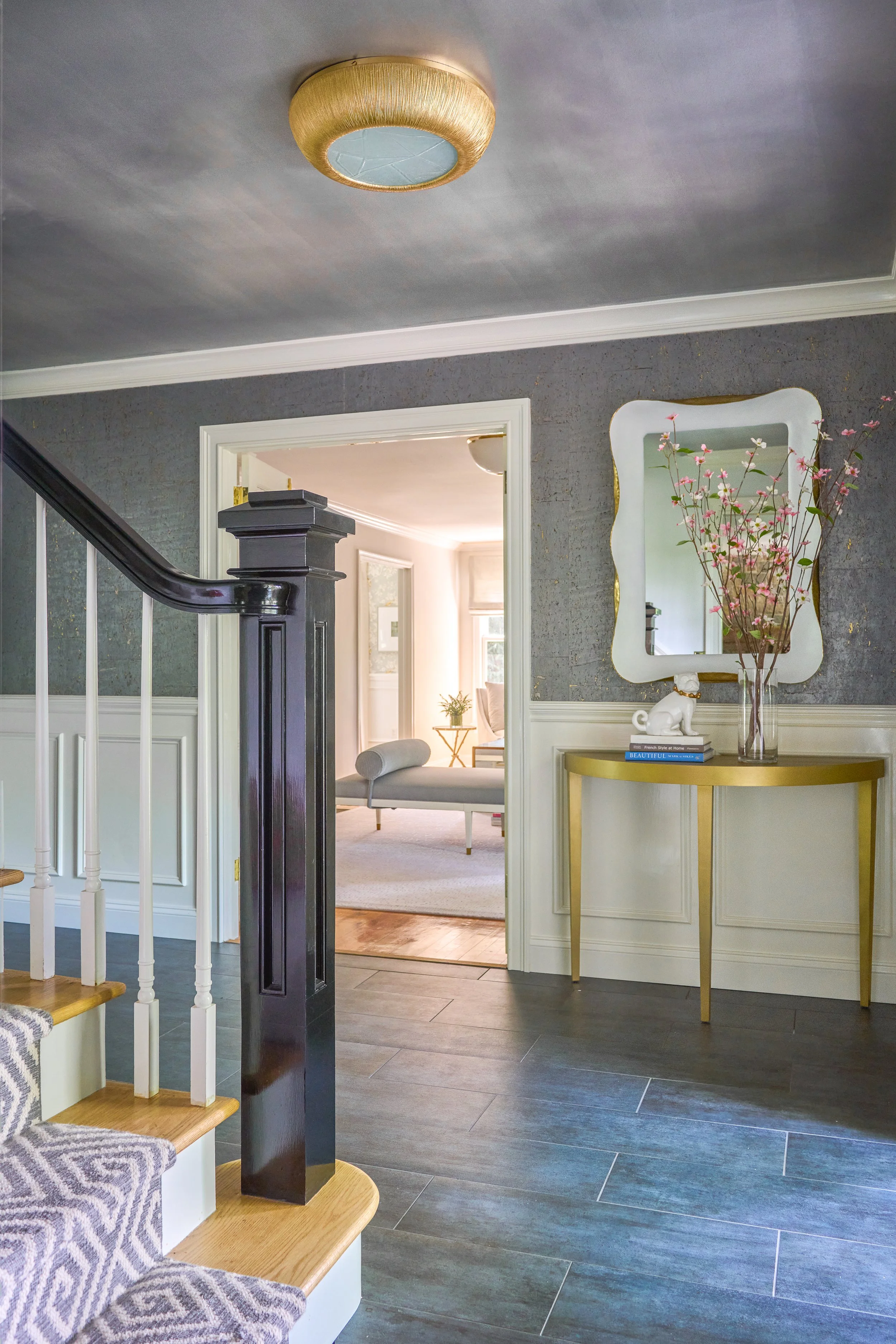 	
Interior of a home featuring a staircase with a navy newell post, a picture of a mirror with a gold frame, pink flowers in a glass vase, and a white elephant figurine on a small table with books, all under a ceiling light.